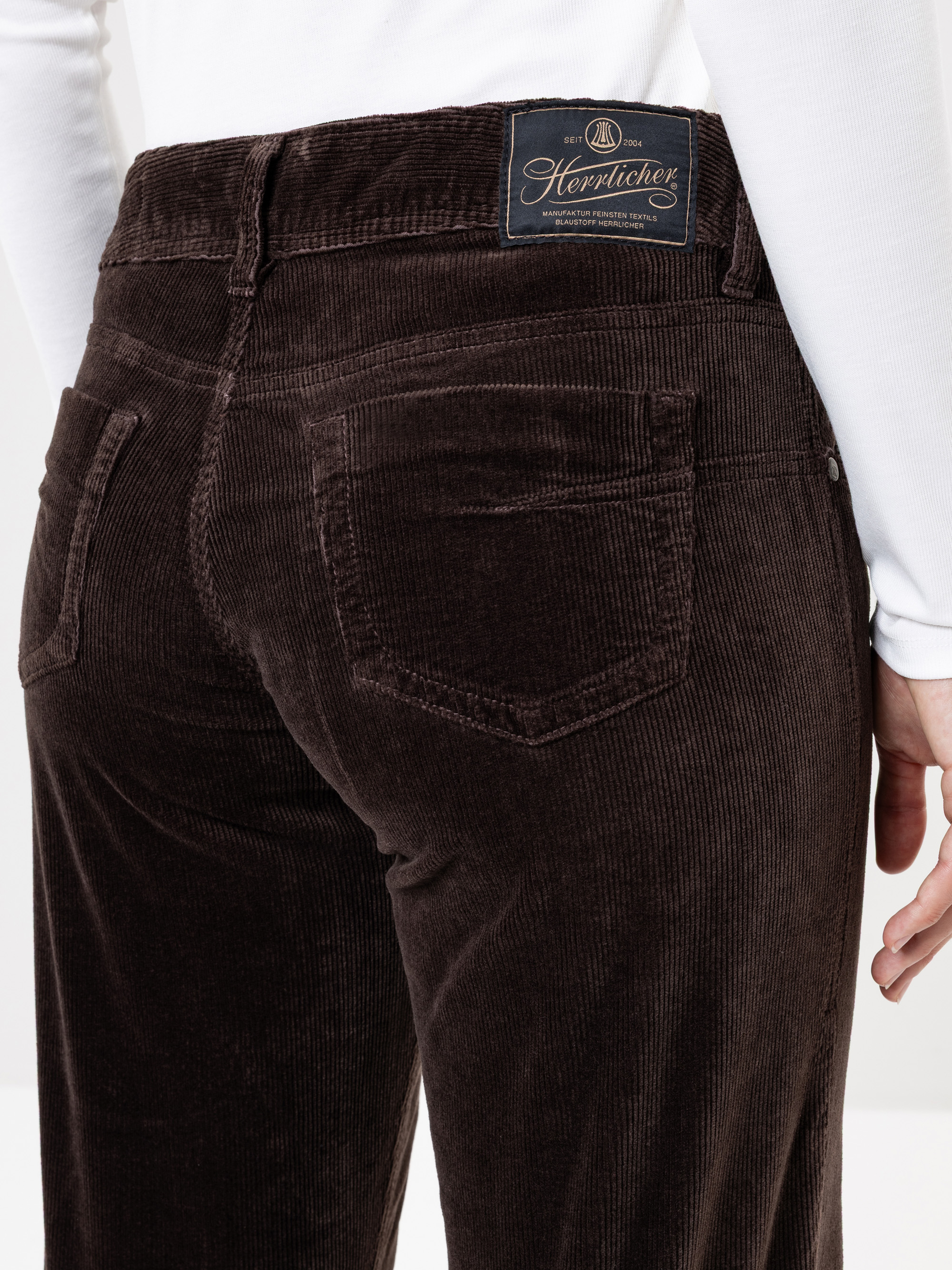 Eine Person, die eine dunkelbraune Cordhose und ein weißes langärmeliges Oberteil trägt, ist von der Taille abwärts abgebildet, mit einer sichtbaren Gesäßtasche und einem dunklen Etikett am Bund.