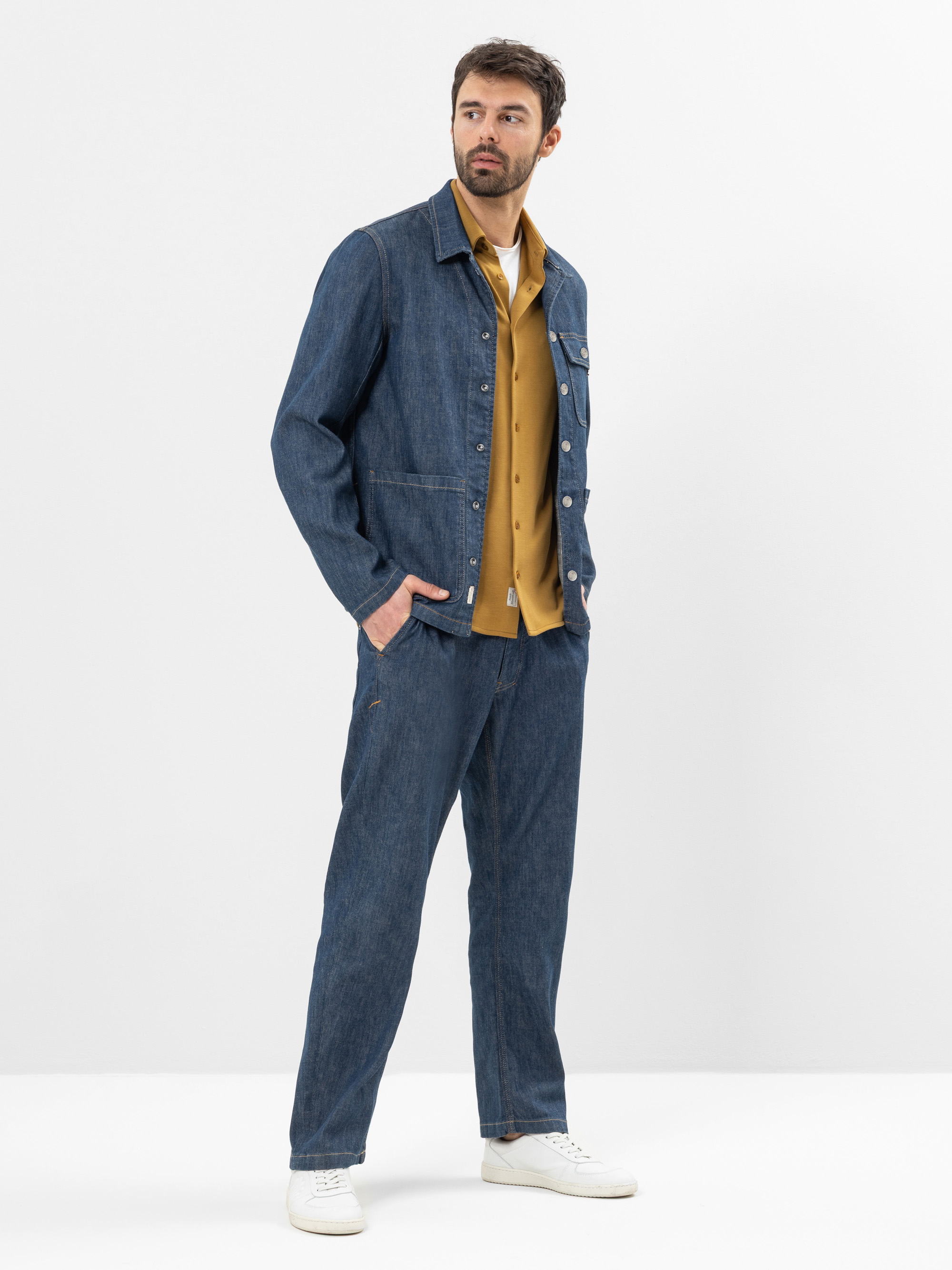 Ein Mann steht vor einem schlichten weißen Hintergrund und trägt eine blaue Jeansjacke und eine dazu passende Jeans, darunter ein senfgelbes Hemd und weiße Turnschuhe. Er hat eine Hand in der Tasche und blickt leicht zur Seite.