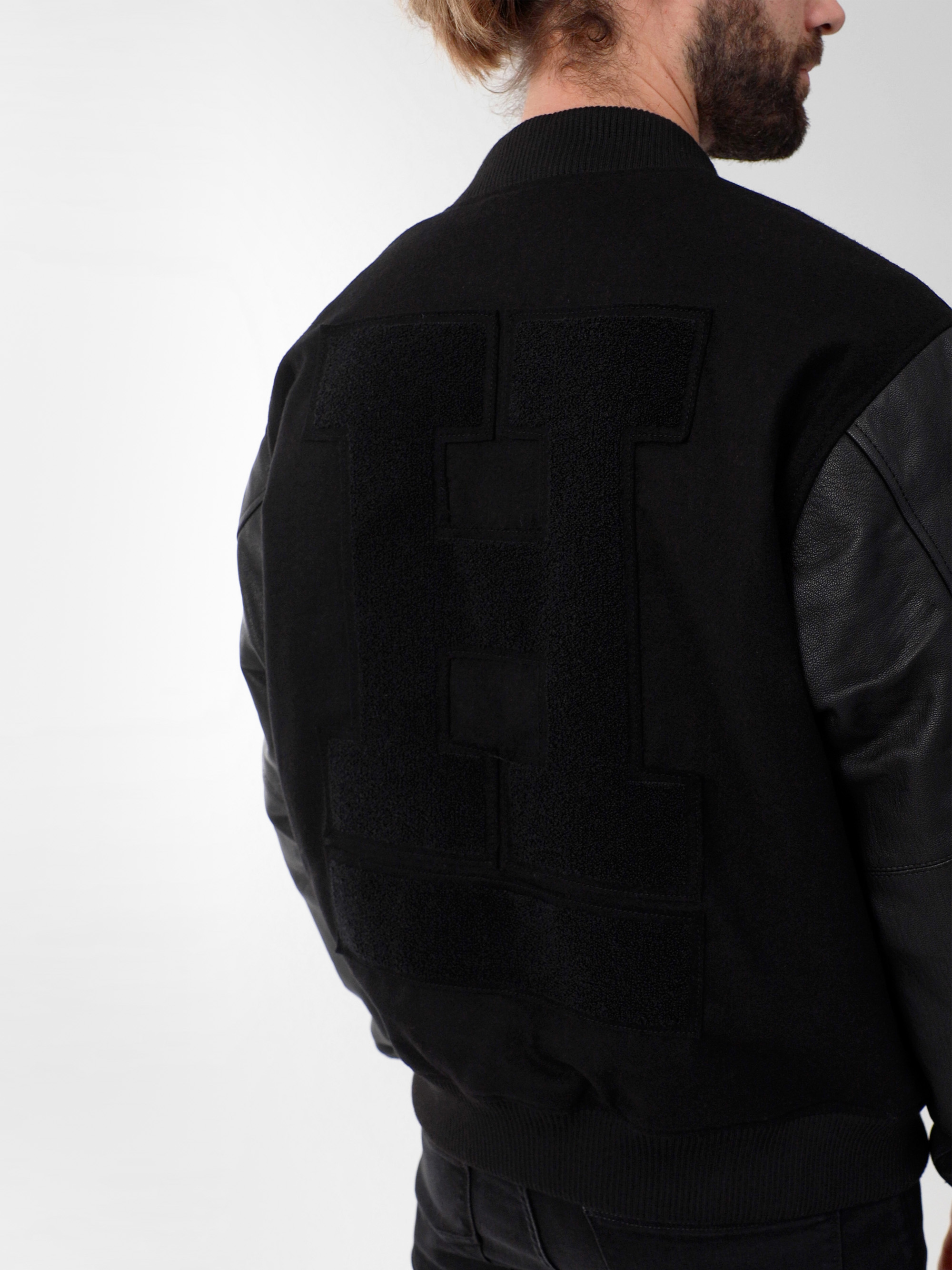 Eine Person mit hellbraunem Haar trägt eine schwarze Sportjacke mit Lederärmeln und einem großen, strukturierten Buchstaben H auf dem Rücken. Das Bild zeigt den Rücken und die Schulter der Person.