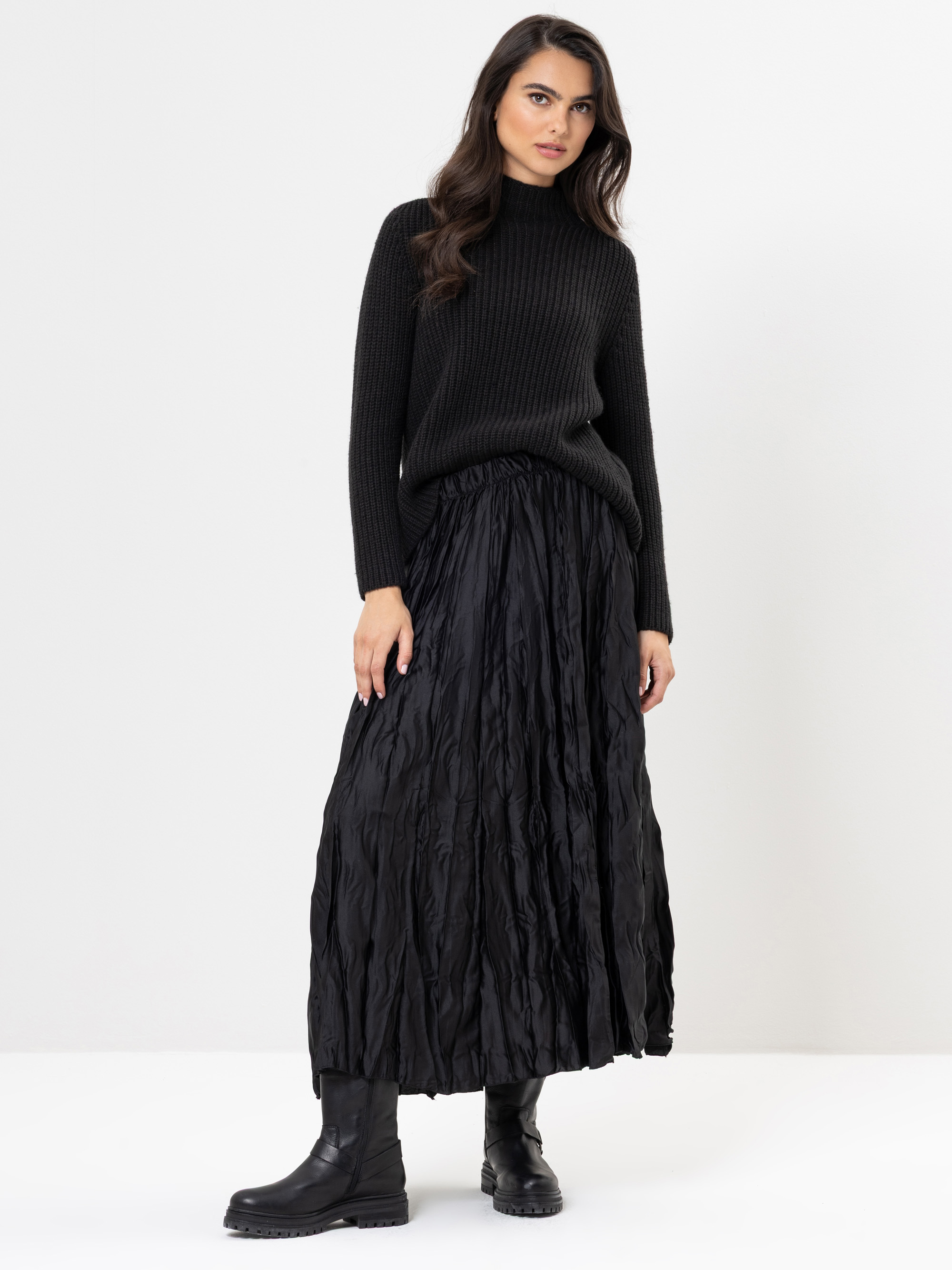Eine Frau steht vor einem weißen Hintergrund und trägt einen schwarzen gerippten Rollkragenpullover, einen langen schwarzen Faltenrock und schwarze Stiefel. Sie hat langes, gewelltes dunkles Haar und einen neutralen Gesichtsausdruck.