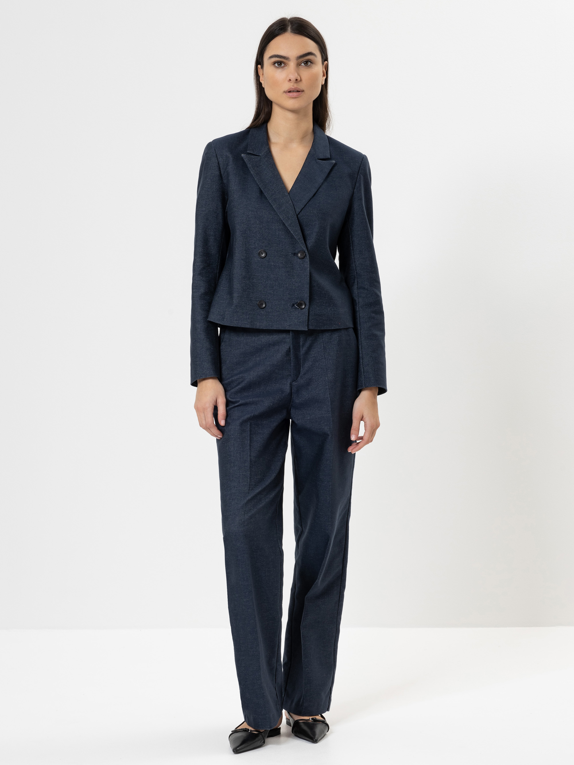 Eine Frau mit langen dunklen Haaren steht vor einem schlichten weißen Hintergrund. Sie trägt einen dunkelblauen zweireihigen Blazer und eine passende Hose mit schwarzen spitzen Schuhen.