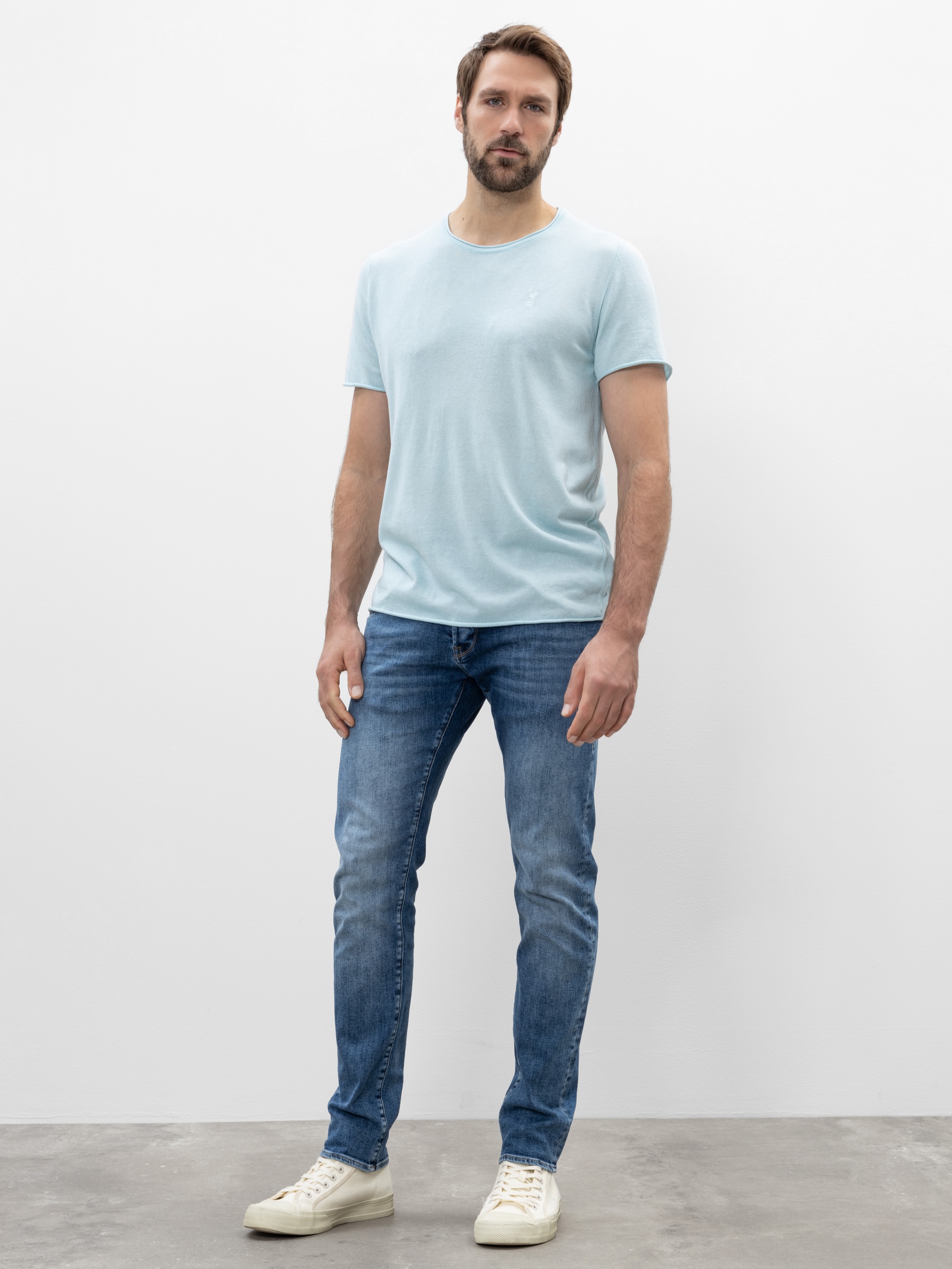 Ein Mann mit kurzen braunen Haaren und einem Bart steht vor einem schlichten weißen Hintergrund und trägt ein hellblaues T-Shirt, blaue Jeans und weiße Turnschuhe. Er hat einen neutralen Gesichtsausdruck und eine entspannte Körperhaltung.