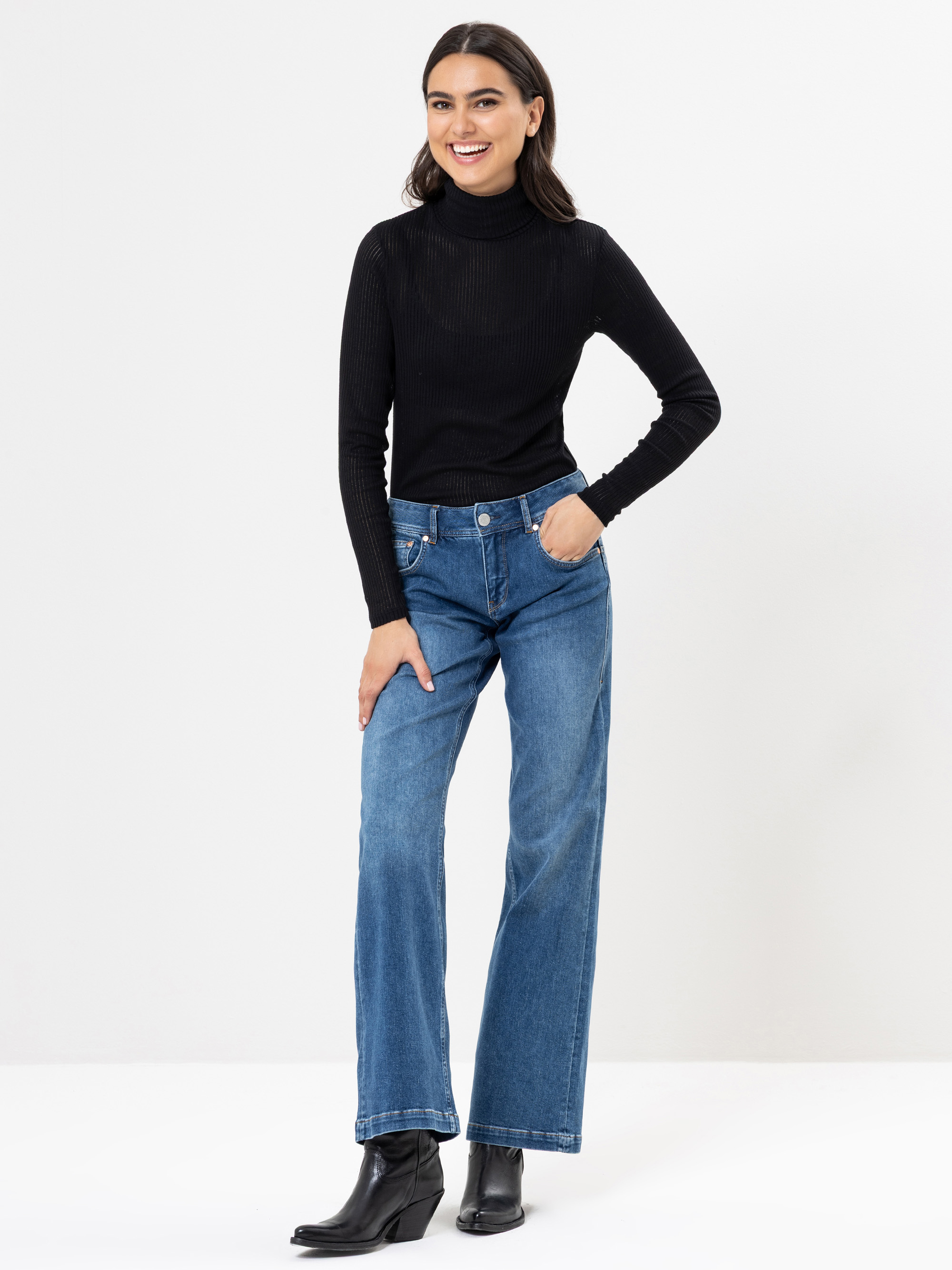 Eine Frau steht lächelnd vor einem weißen Hintergrund. Sie trägt einen schwarzen Rollkragenpullover, blaue Jeans mit weitem Bein und schwarze Ankle Boots. Eine Hand ruht lässig auf ihrem Knie.