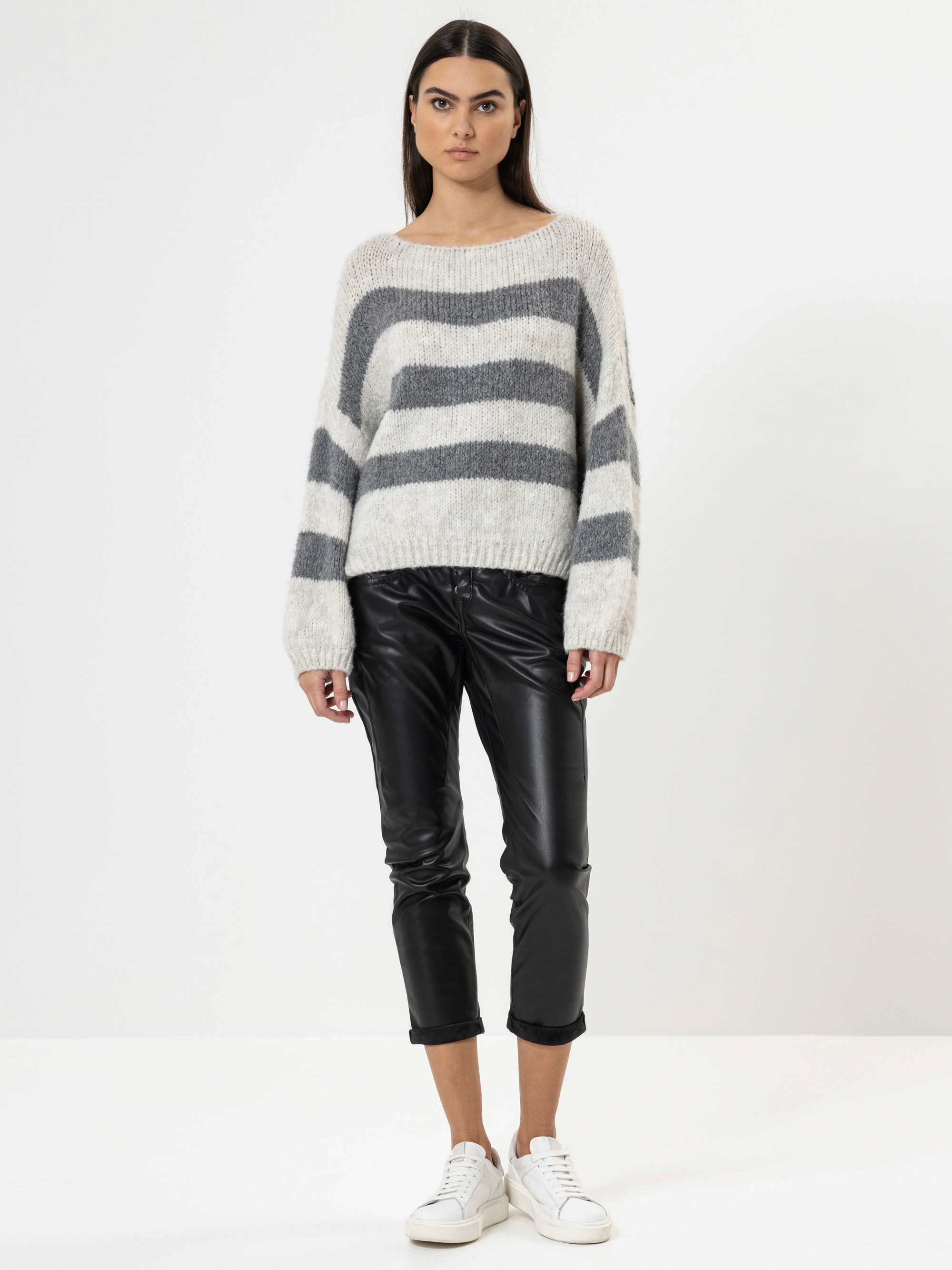 Eine Frau steht vor einem schlichten weißen Hintergrund und trägt einen cremefarbenen Pullover mit breiten grauen Streifen, eine schwarze, kurze Lederhose und weiße Turnschuhe. Sie schaut mit neutralem Blick geradeaus.