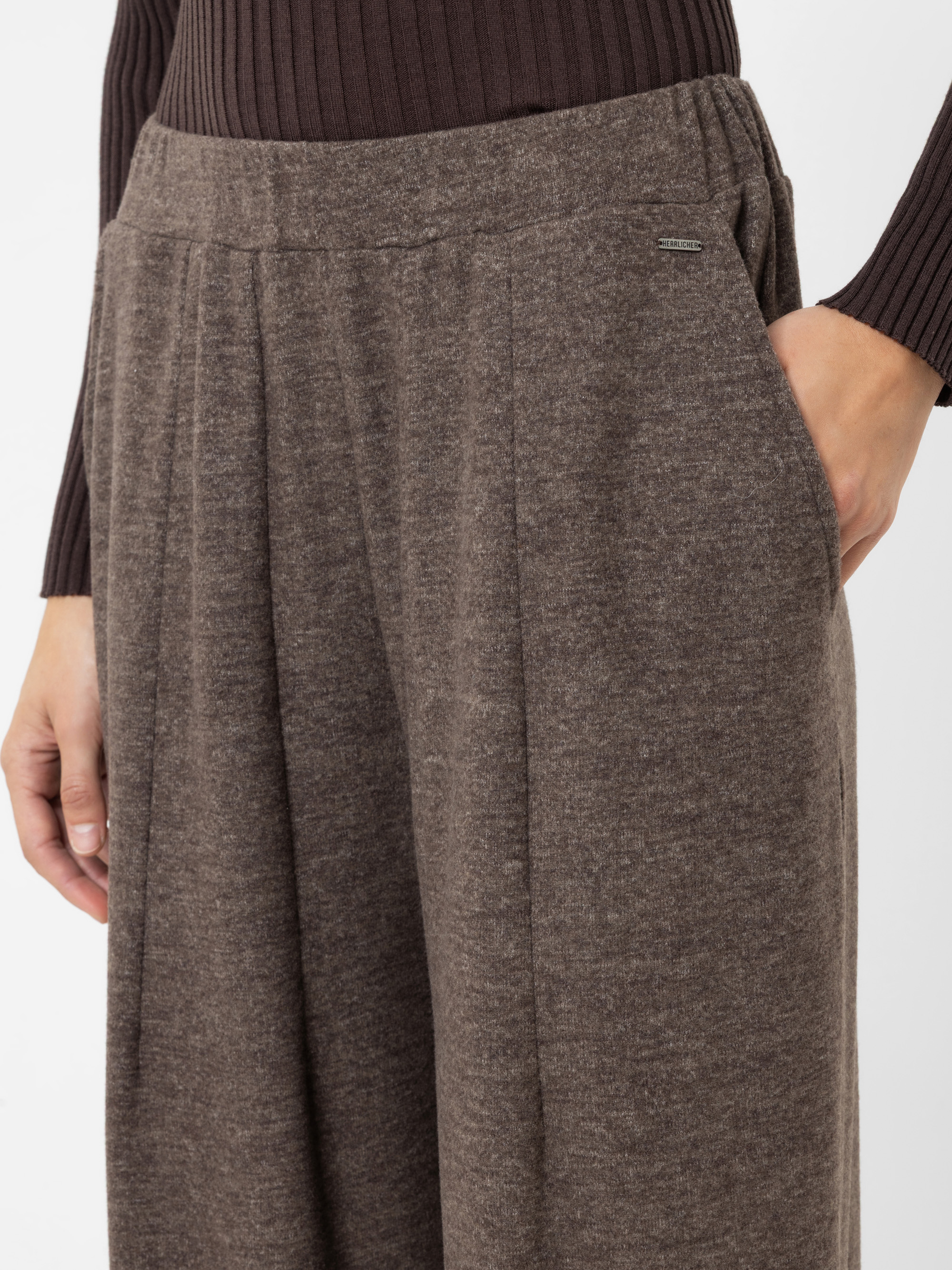 Eine Person, die eine locker sitzende braune Hose trägt, die rechte Hand in der Tasche hat und ein geripptes, dunkelbraunes Langarm-Oberteil anhat. Der Fokus liegt auf dem Hosenbund und dem Oberschenkelbereich.