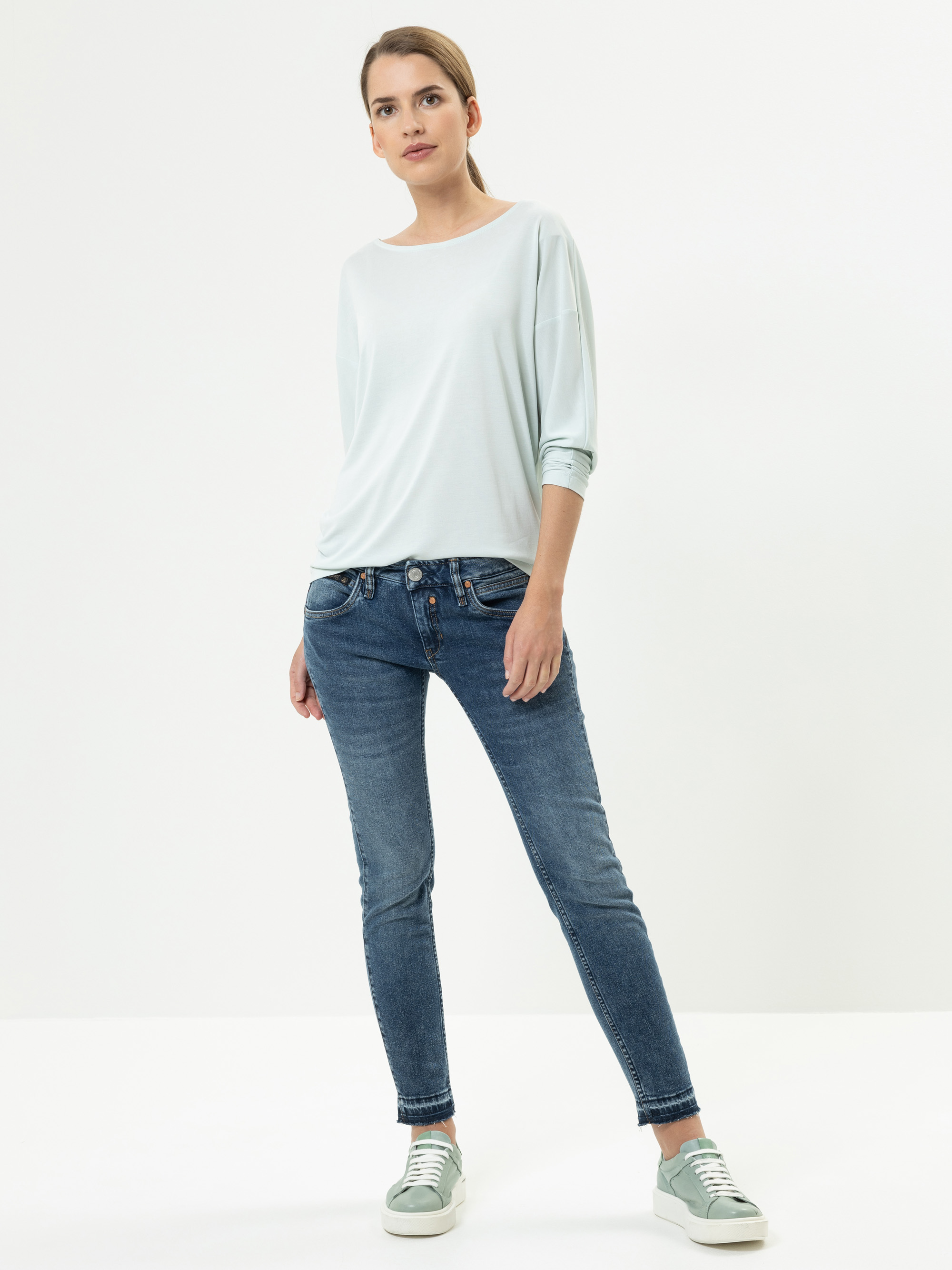 Eine Frau steht vor einem schlichten weißen Hintergrund und trägt ein lockeres hellblaues, langärmeliges Oberteil, blaue Skinny Jeans und hellgrüne Turnschuhe. Sie hat die Hände in den Taschen und blickt leicht zur Seite.