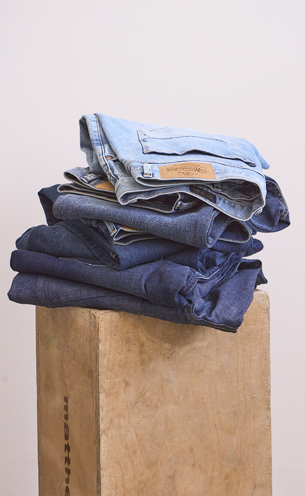 Auf einer Holzkiste vor einem schlichten, hellen Hintergrund liegt ein Stapel aus drei Paaren gefalteter Bluejeans in verschiedenen Farbtönen, von hell bis dunkel.