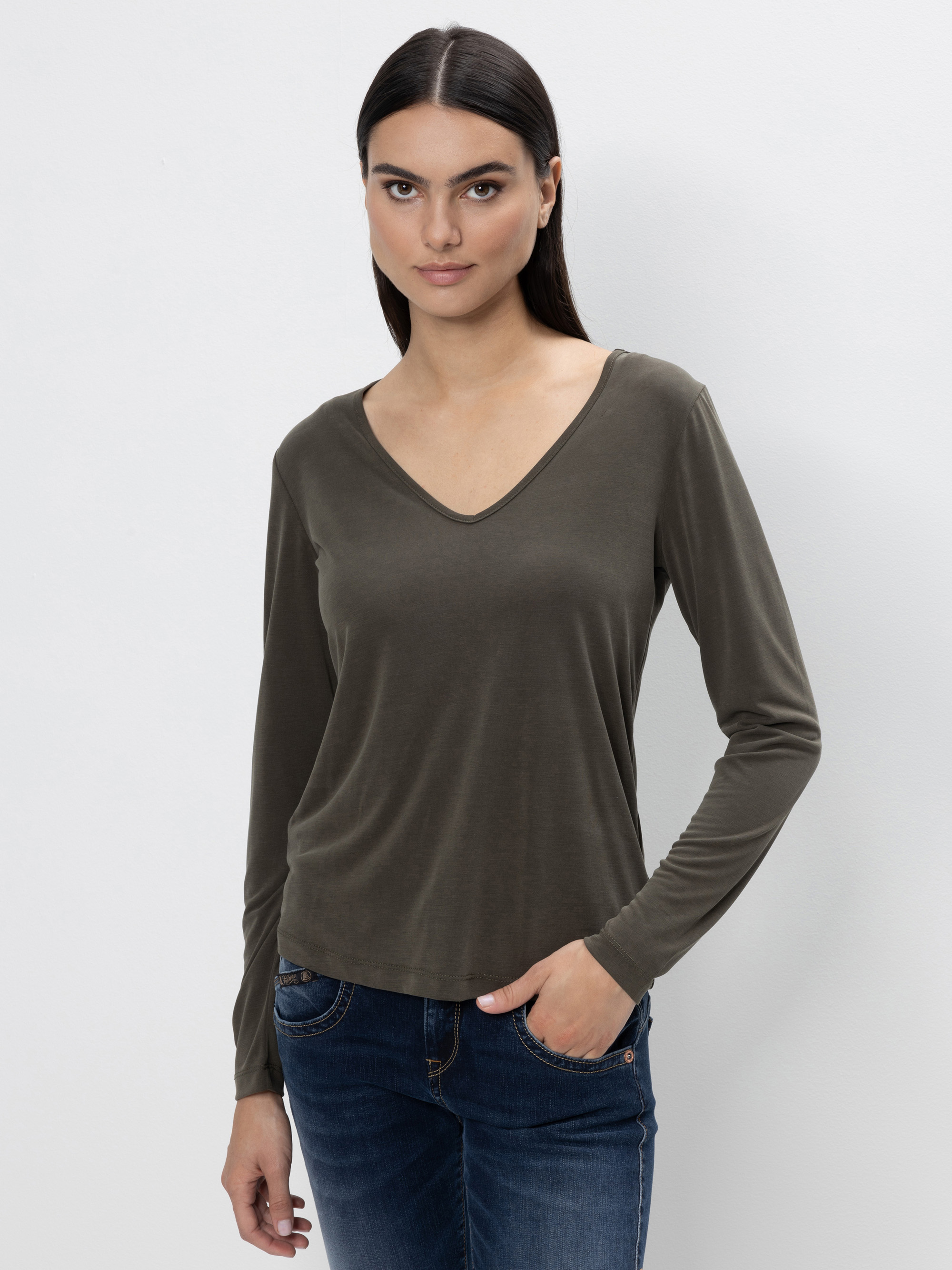 Eine Frau mit glattem dunklem Haar, die ein langärmeliges olivgrünes Oberteil mit V-Ausschnitt und blaue Jeans trägt, steht vor einem schlichten hellen Hintergrund und hat eine Hand in der Tasche.