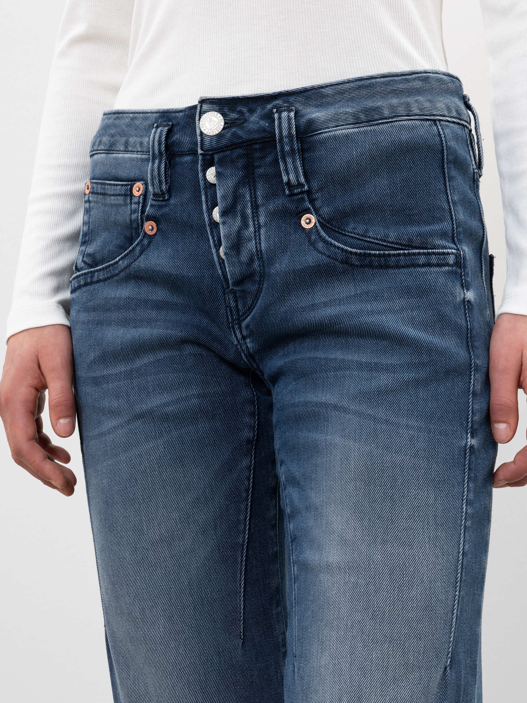 Eine Person trägt ein weißes langärmeliges Oberteil und eine blaue Jeans mit hoher Taille und Knopfleiste, die von der Taille bis zur Mitte des Oberschenkels reicht. Die Jeans sieht verblasst und leicht abgenutzt aus.