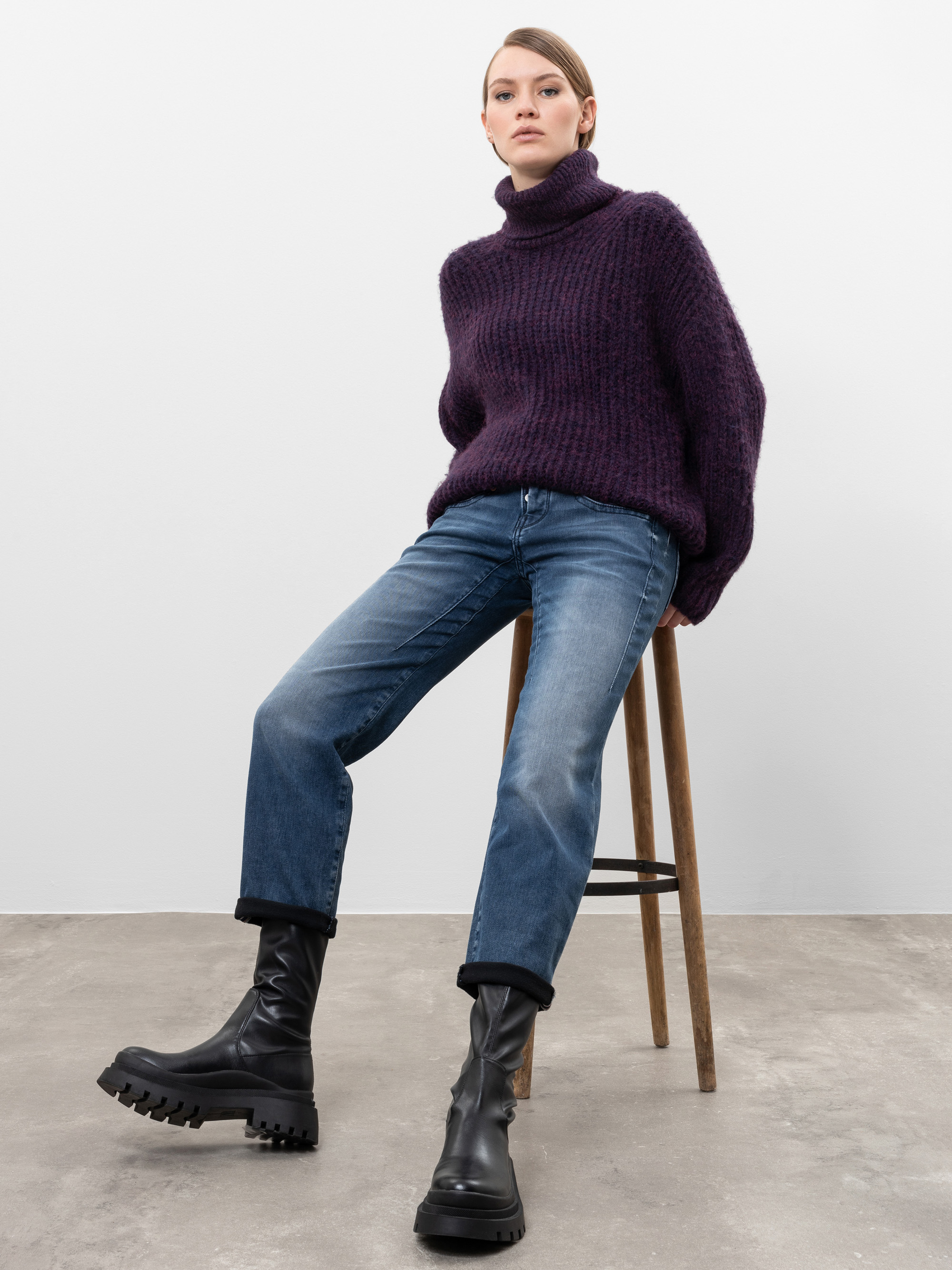 Eine Person in einem dicken lila Rollkragenpullover, blauen Jeans und schwarzen Stiefeln sitzt auf einem Holzhocker vor einer schlichten weißen Wand und einem Betonboden.