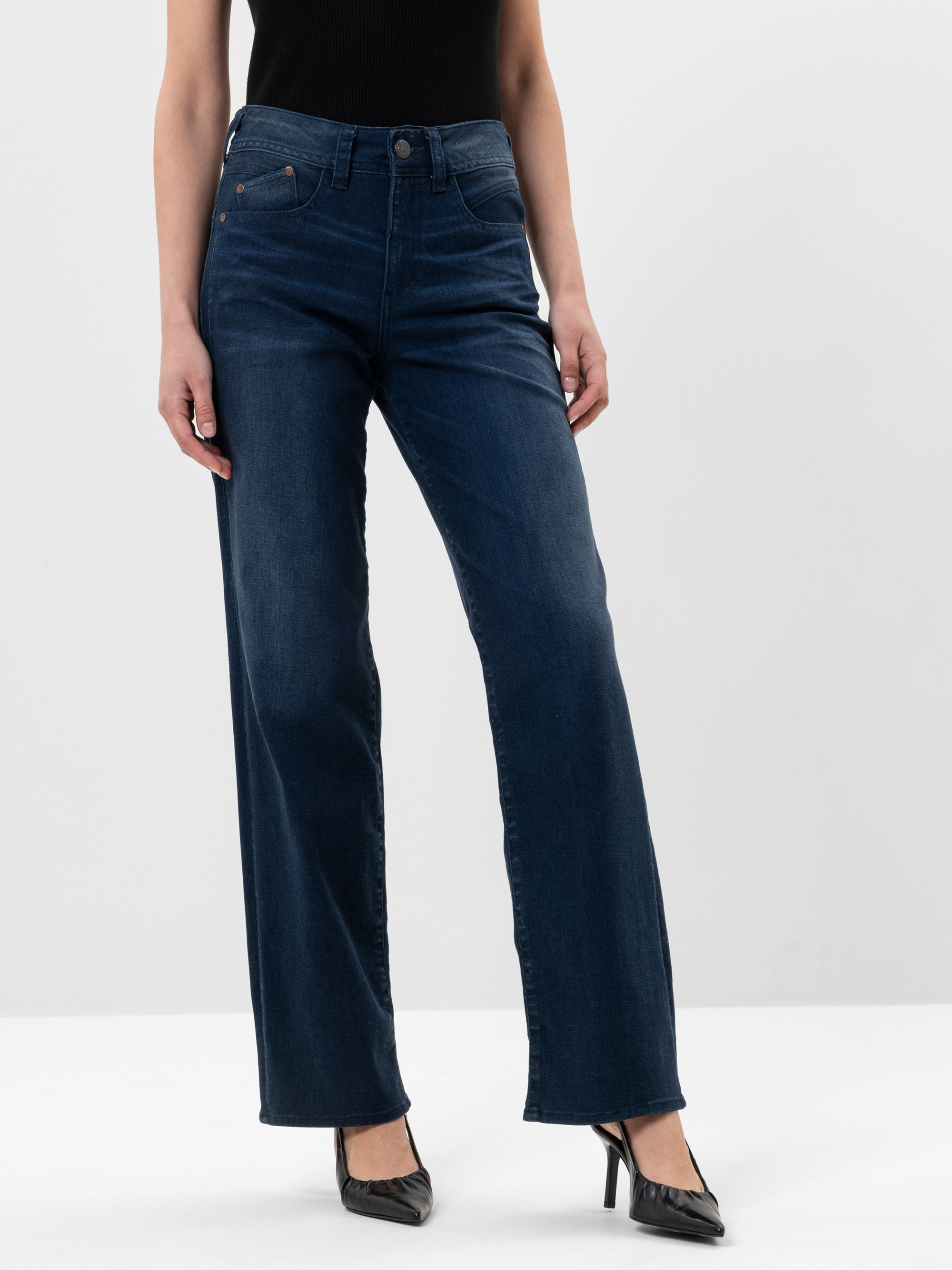 Eine Person, die ein schwarzes ärmelloses Oberteil, dunkelblaue Jeans mit weitem Bein und schwarze Schuhe mit Absätzen trägt, steht vor einem schlichten weißen Hintergrund.