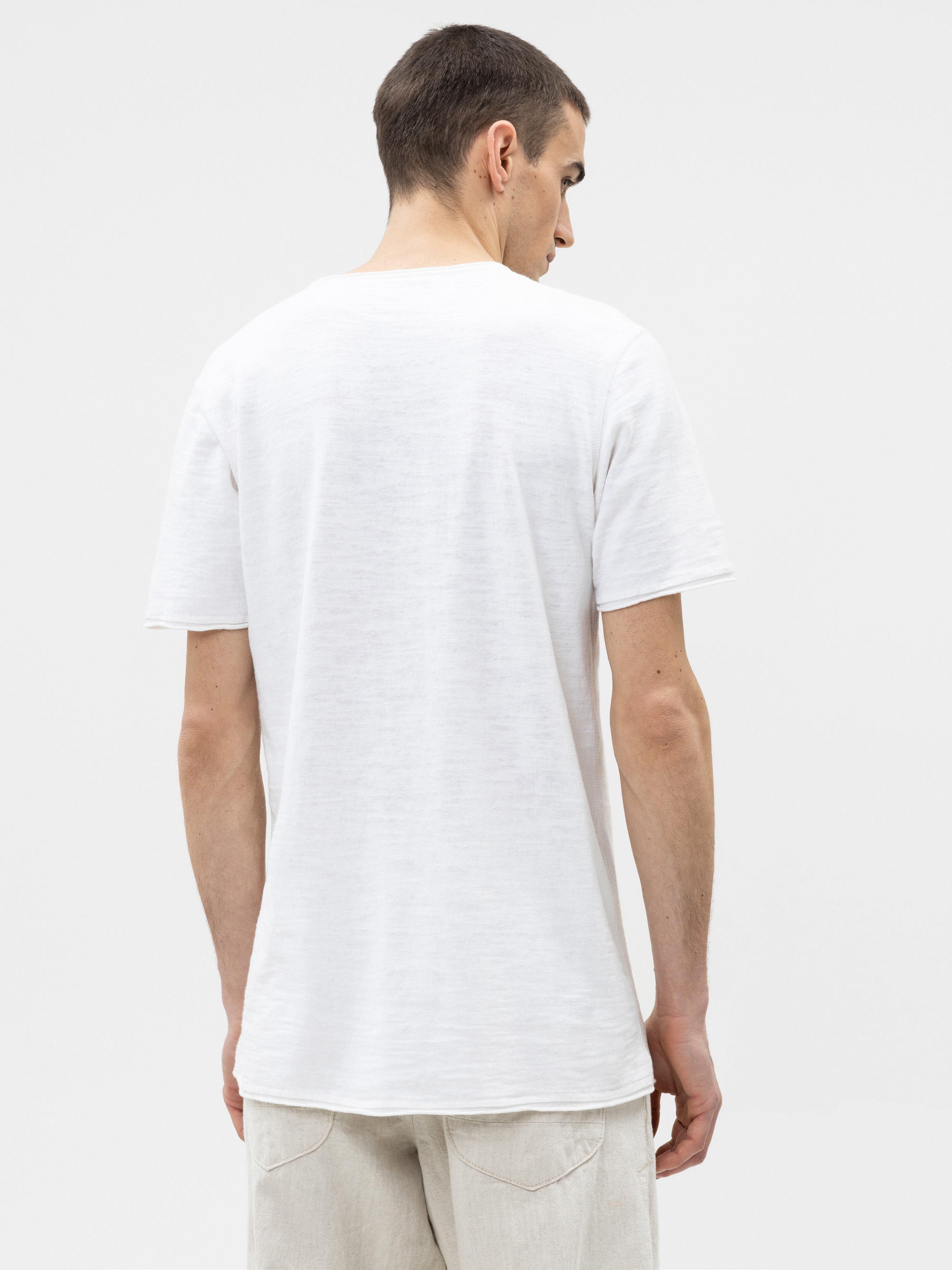 Ein Mann mit kurzen braunen Haaren steht und schaut von der Kamera weg. Er trägt ein schlichtes weißes kurzärmeliges T-Shirt und eine hellbeige Hose vor einem einfarbig weißen Hintergrund.