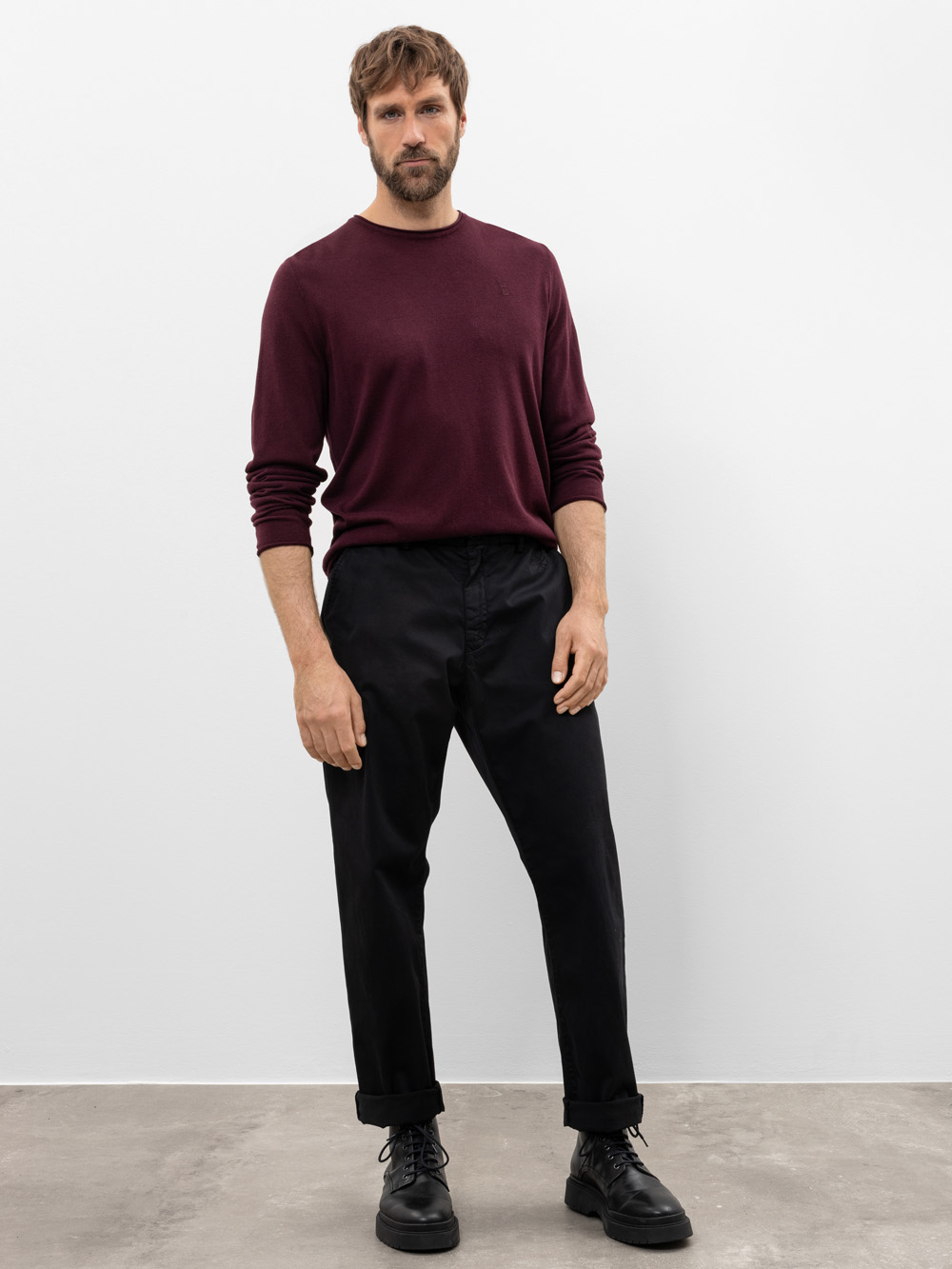 Ein Mann mit kurzen braunen Haaren und einem Bart steht vor einer schlichten weißen Wand. Er trägt einen langärmeligen weinroten Pullover, eine schwarze Hose und schwarze Schuhe. Sein Gesichtsausdruck ist neutral und seine Hände ruhen an den Seiten.