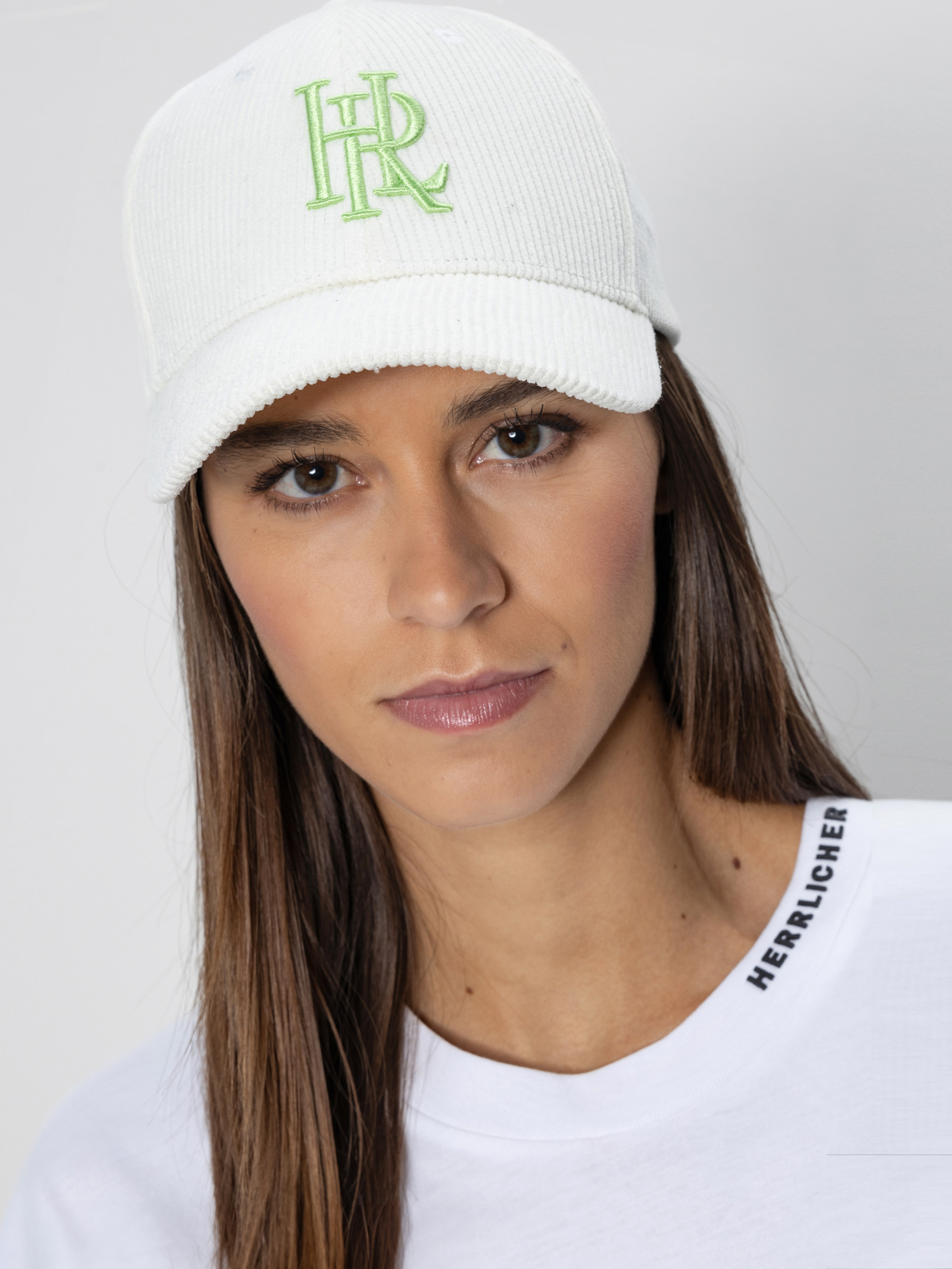 Eine Person mit langen braunen Haaren trägt eine weiße Mütze mit grüner Stickerei und ein weißes Hemd mit dem Aufdruck HERALUCHER am Kragen. Der Hintergrund ist einfarbig und hell gefärbt.