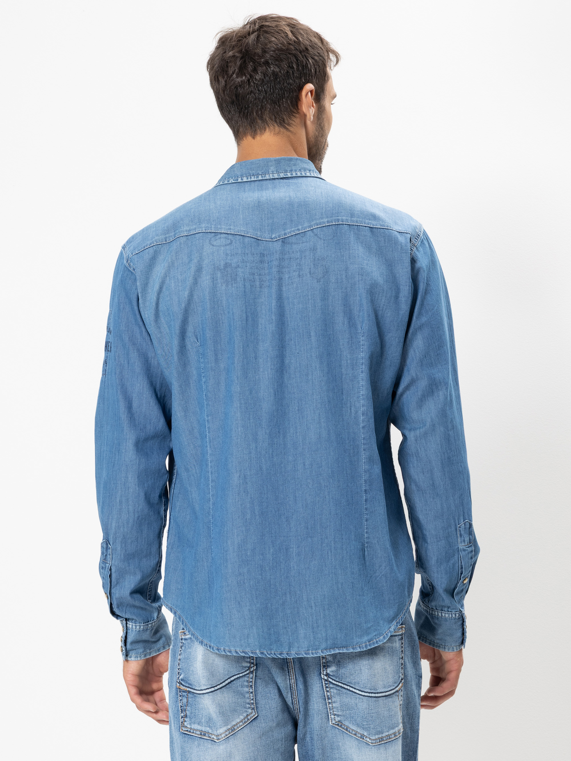 Ein Mann steht mit dem Rücken zur Kamera, trägt ein langärmeliges Jeanshemd und hellblaue Jeans vor einem schlichten weißen Hintergrund.