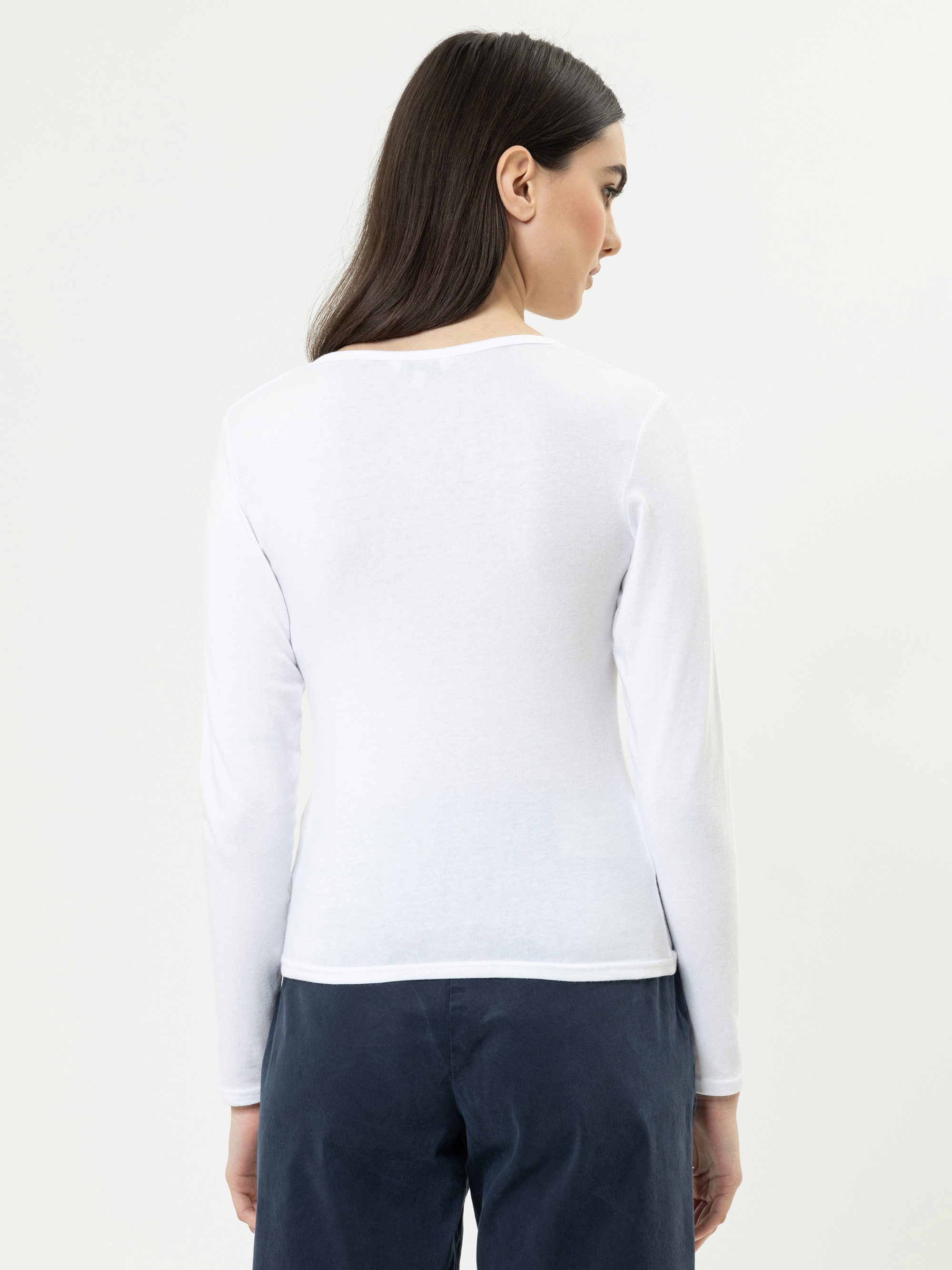 Eine Frau mit langen dunklen Haaren wird von hinten gezeigt. Sie trägt ein weißes, langärmeliges Oberteil und eine dunkle Hose und steht vor einem schlichten, hellen Hintergrund.