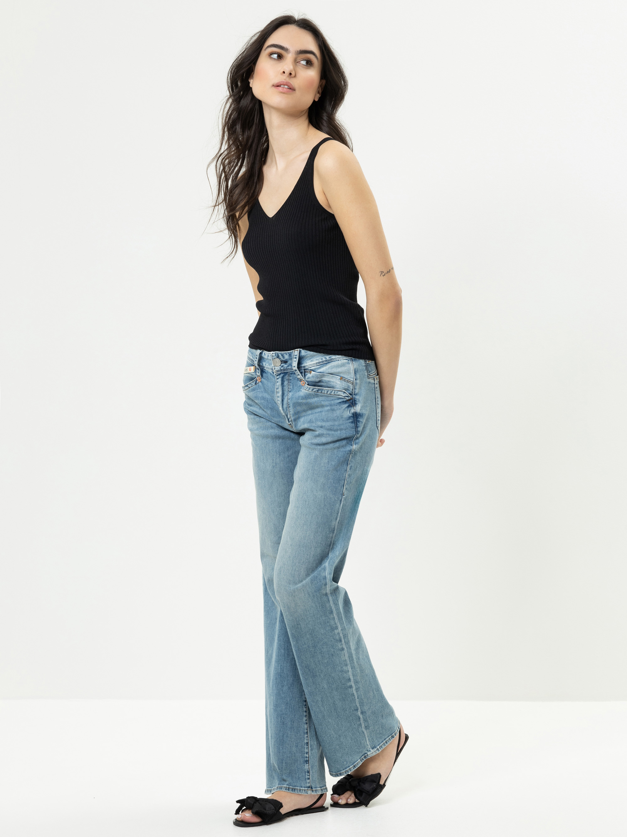 Eine Frau mit langen dunklen Haaren, die ein schwarzes ärmelloses Oberteil, hellblaue Jeans und schwarze flache Schuhe mit Schleife trägt, steht vor einem schlichten weißen Hintergrund und blickt mit entspannter Haltung leicht zur Seite.