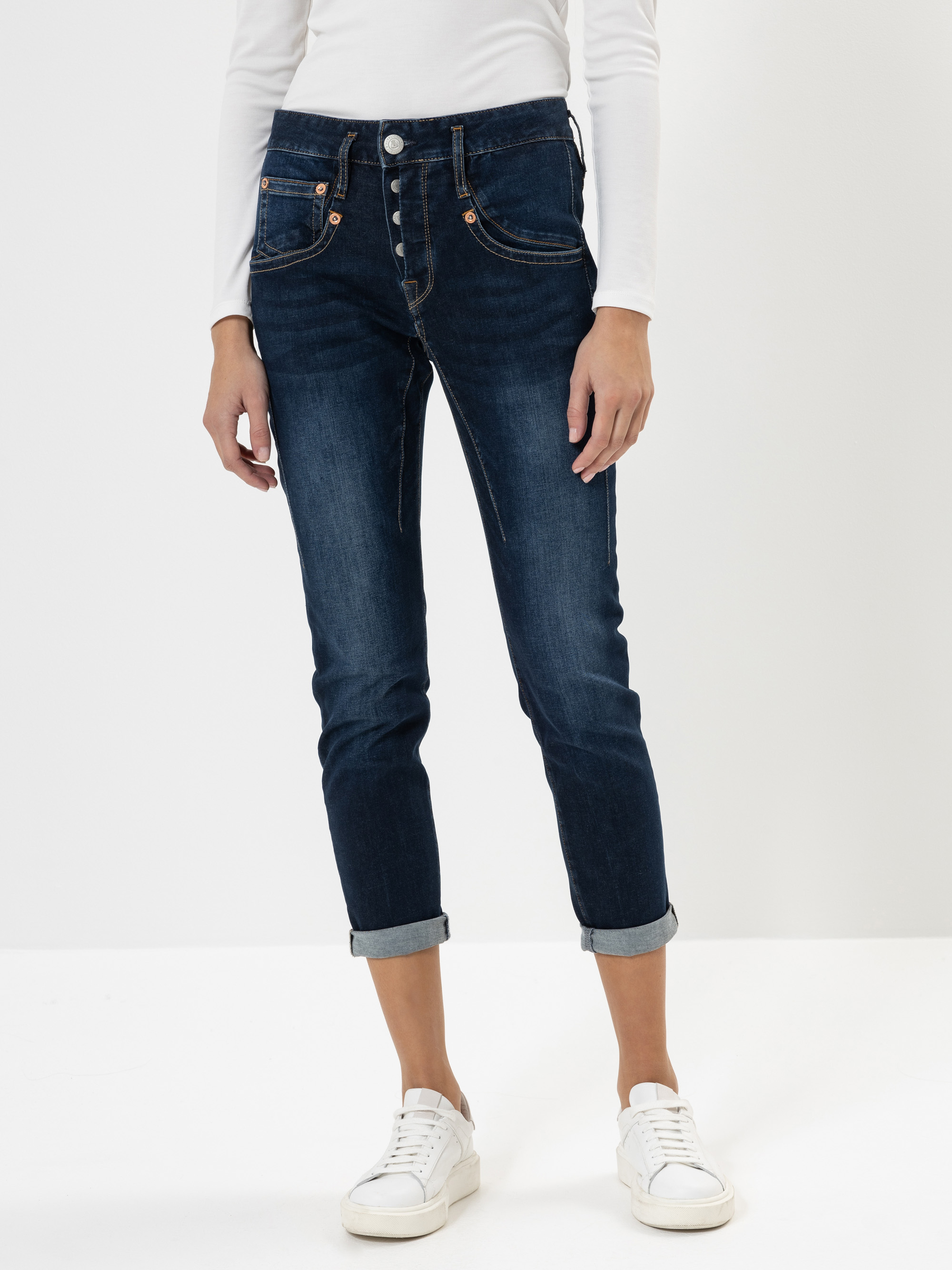 Eine Person, die eine dunkelblaue Jeans mit Bündchen, ein langärmeliges weißes Oberteil und weiße Turnschuhe trägt, steht vor einem schlichten weißen Hintergrund. Ihre Hände liegen entspannt an der Seite.