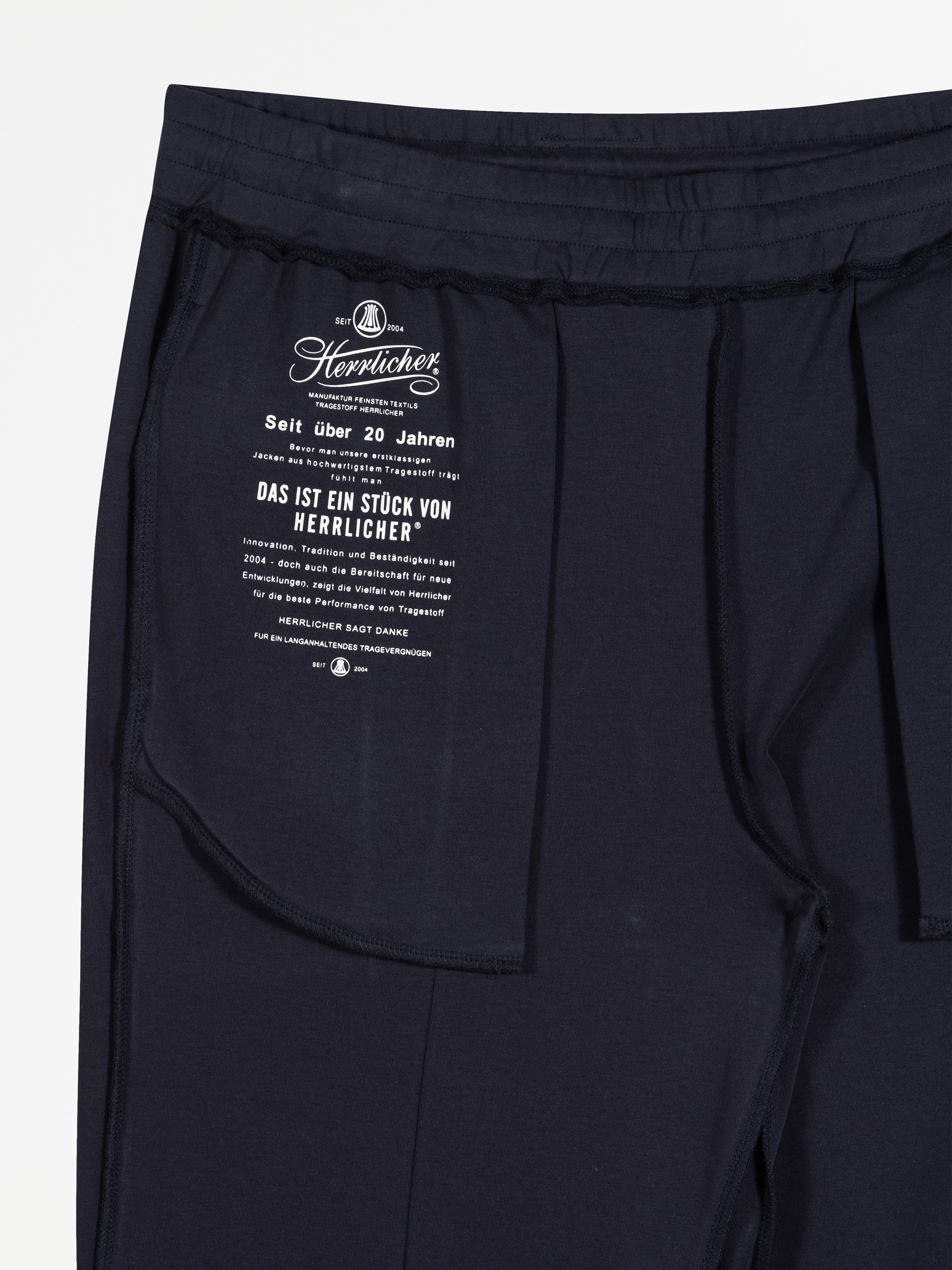 Marineblaue Jogginghose mit einem weiß bedruckten Etikett am Innenbund, auf dem Markenzeichen und Pflegehinweise in deutscher Sprache stehen. Die Hose liegt flach auf einer weißen Unterlage.