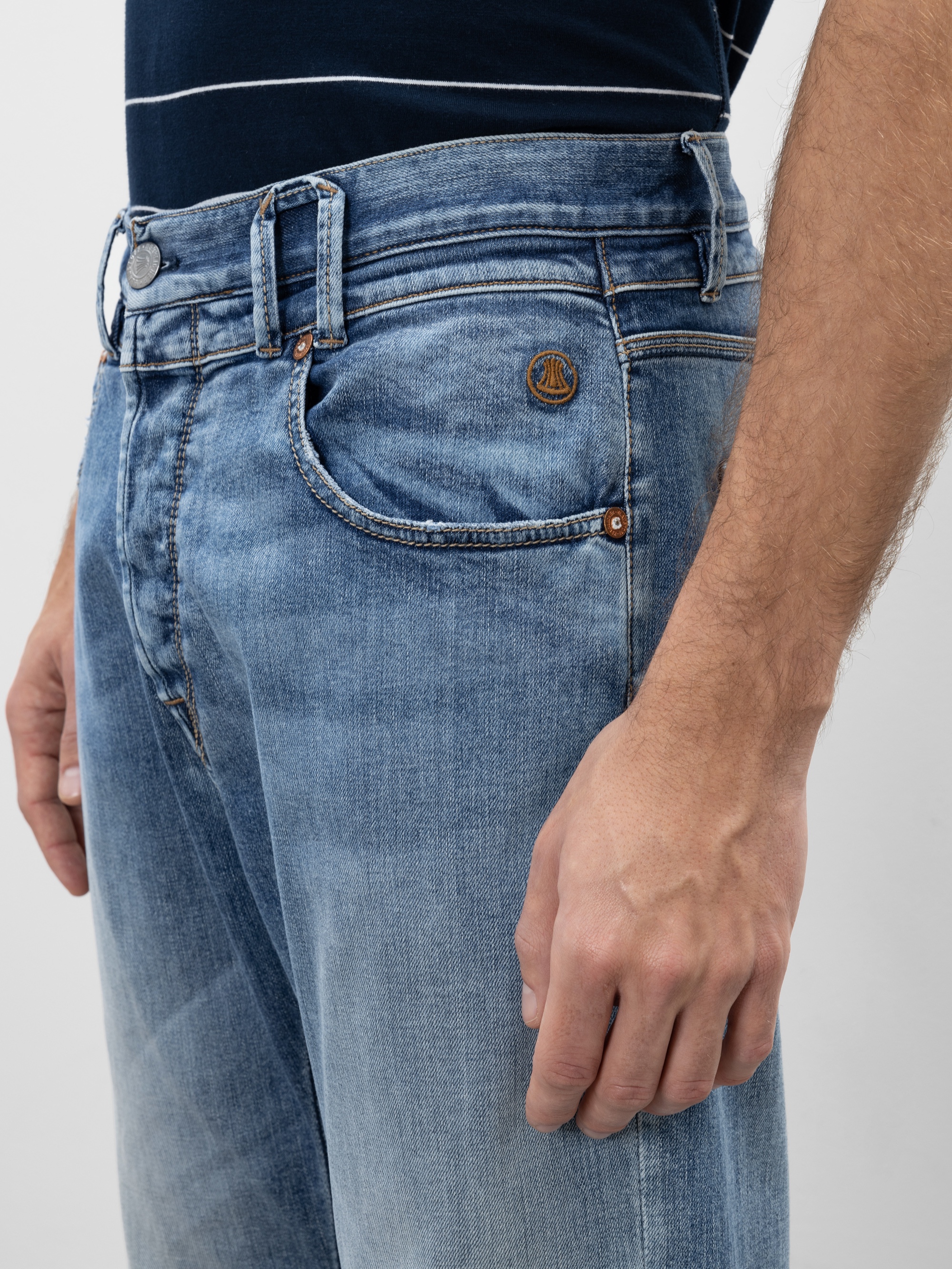 Eine Person trägt eine hellblaue Jeans und ein dunkles Hemd. Das Bild konzentriert sich auf den Bund und den Taschenbereich der Jeans und zeigt ein kleines Logo auf der Tasche und die rechte Hand der Person, die an ihrer Seite ruht.