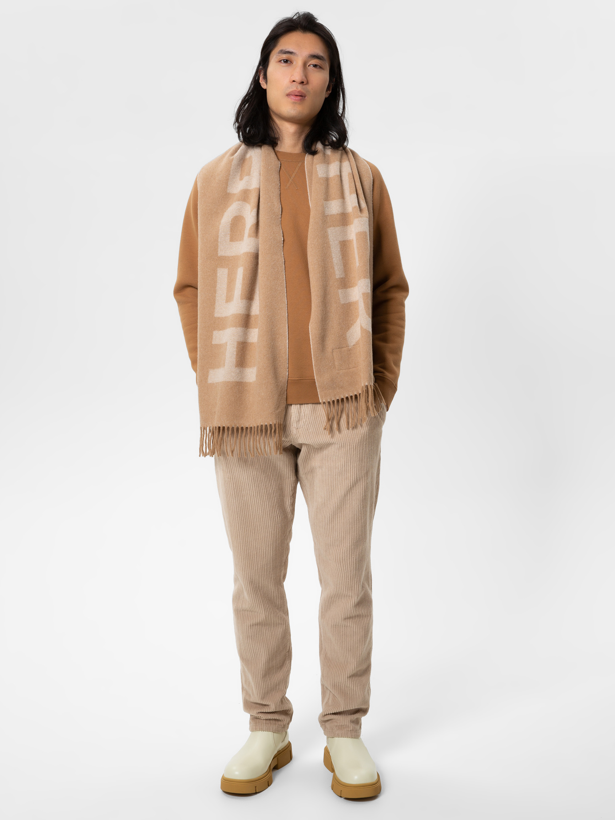 Eine Person mit langen dunklen Haaren steht mit dem Gesicht nach vorne und trägt einen hellbraunen Schal mit Fransen, ein braunes Sweatshirt, eine beigefarbene Cordhose und helle Stiefel vor einem schlichten weißen Hintergrund.