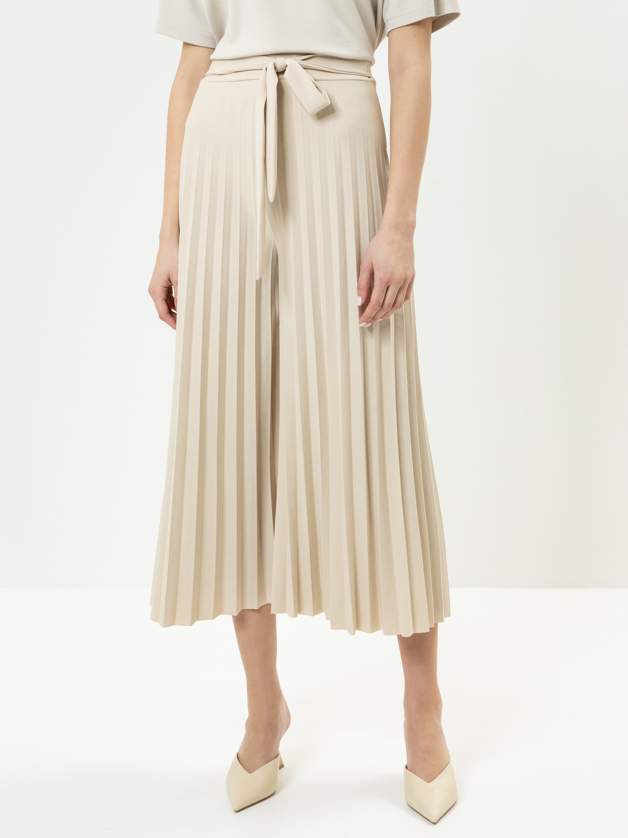 Eine Person, die einen beigen plissierten Midirock mit passendem Bindegürtel, ein leichtes kurzärmeliges Oberteil und spitze beigefarbene Schuhe trägt, steht vor einem schlichten weißen Hintergrund.