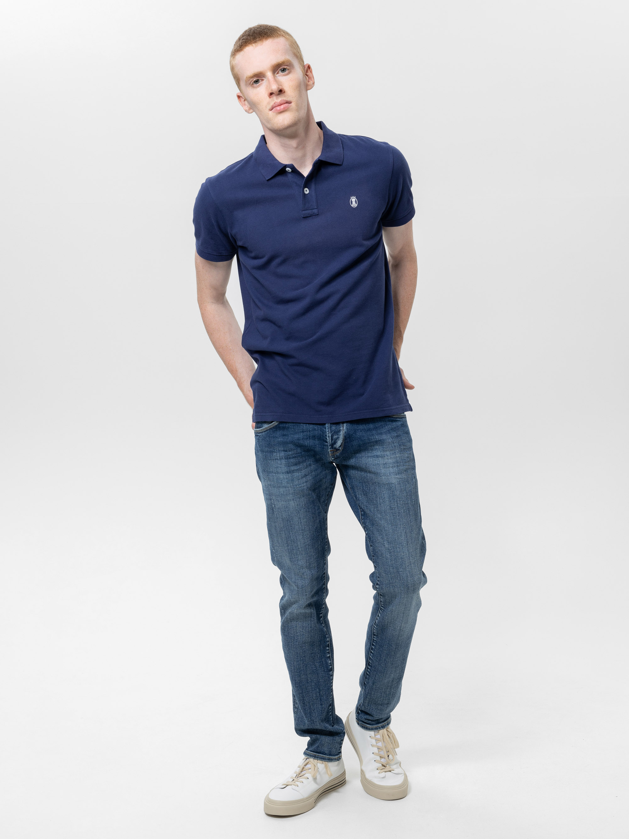 Ein Mann mit kurzen roten Haaren, der ein marineblaues Poloshirt, blaue Jeans und weiße Turnschuhe trägt, steht mit den Händen auf dem Rücken vor einem schlichten weißen Hintergrund.