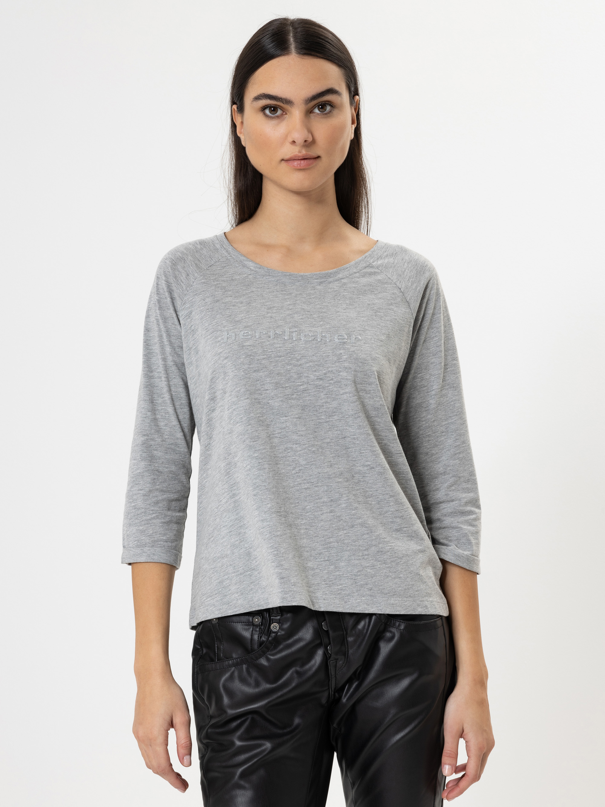 Eine Frau mit langen dunklen Haaren, die ein hellgraues, dreiviertelärmliges Oberteil und eine schwarze Lederhose trägt, steht vor einem schlichten weißen Hintergrund und schaut mit neutralem Blick direkt in die Kamera.