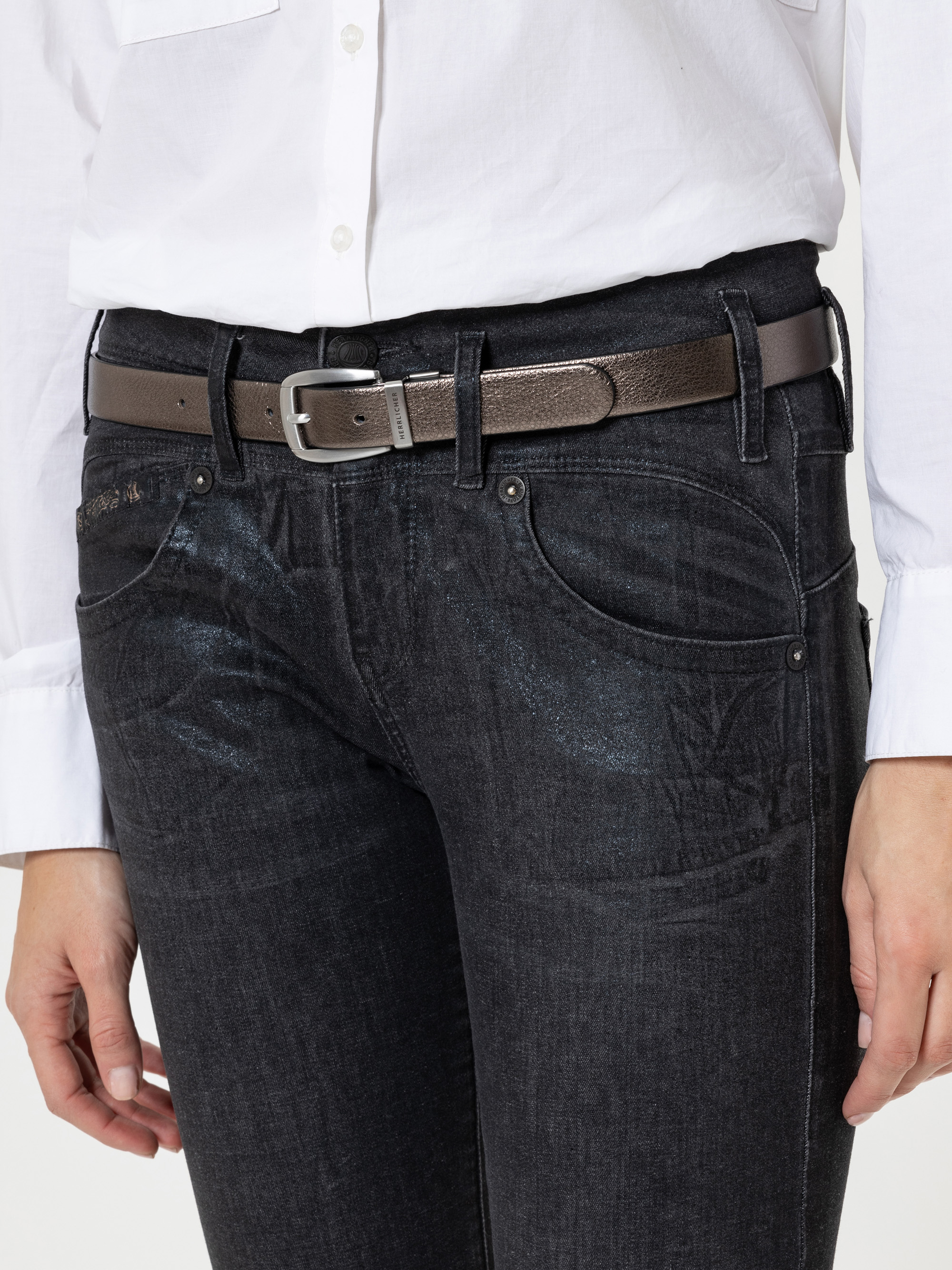 Eine Person, die ein weißes Button-up-Hemd trägt, das in eine dunkle Jeans gesteckt ist, die mit einem braunen Ledergürtel mit silberner Schnalle geschlossen ist. Nur der Oberkörper und die Oberschenkel sind sichtbar.