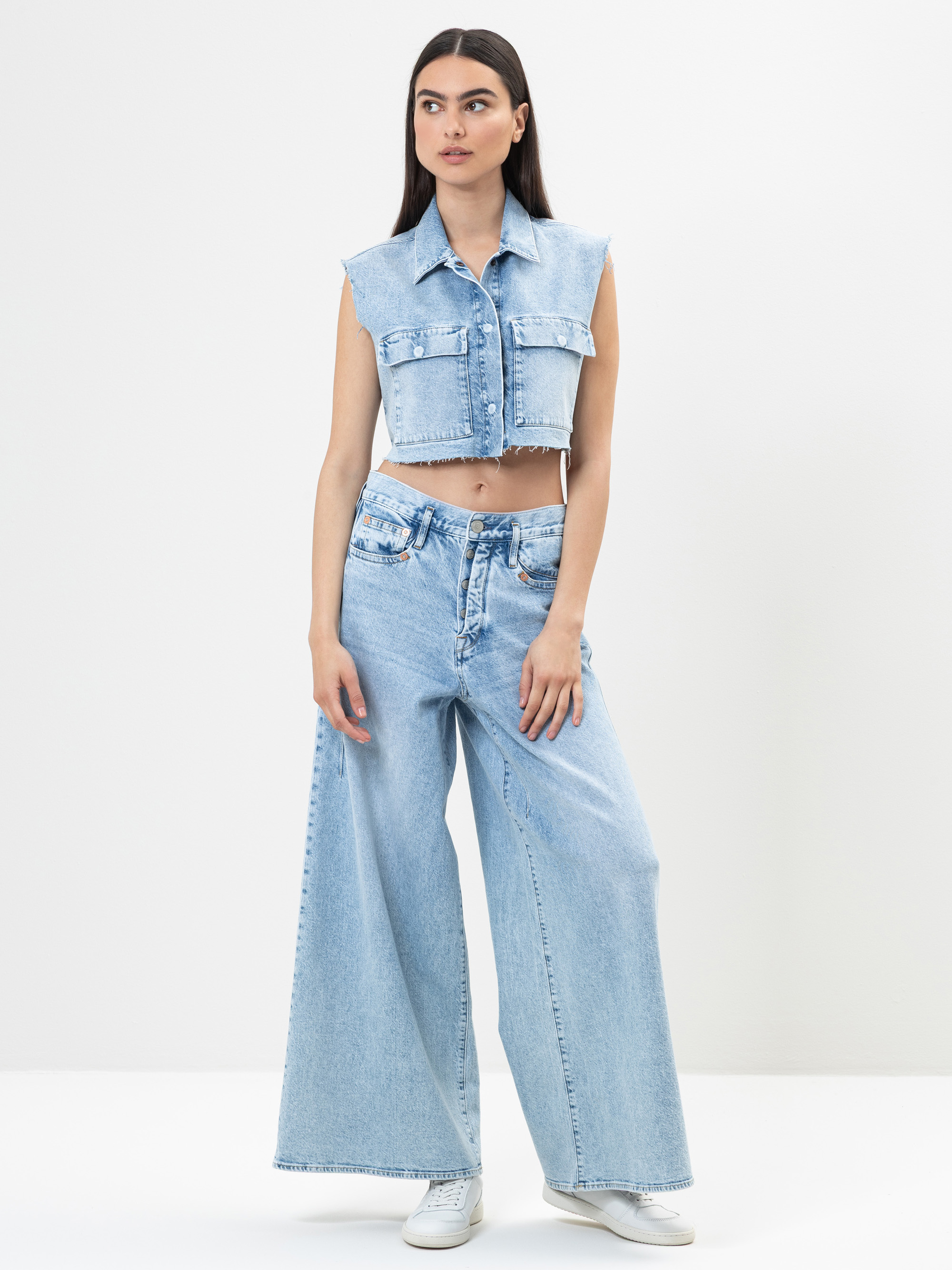 Eine Frau steht vor einem schlichten weißen Hintergrund und trägt eine ärmellose, kurz geschnittene Jeansweste und eine hellblaue Jeans mit weitem Bein und hoher Taille, kombiniert mit weißen Turnschuhen. Sie hat langes glattes Haar und einen neutrale