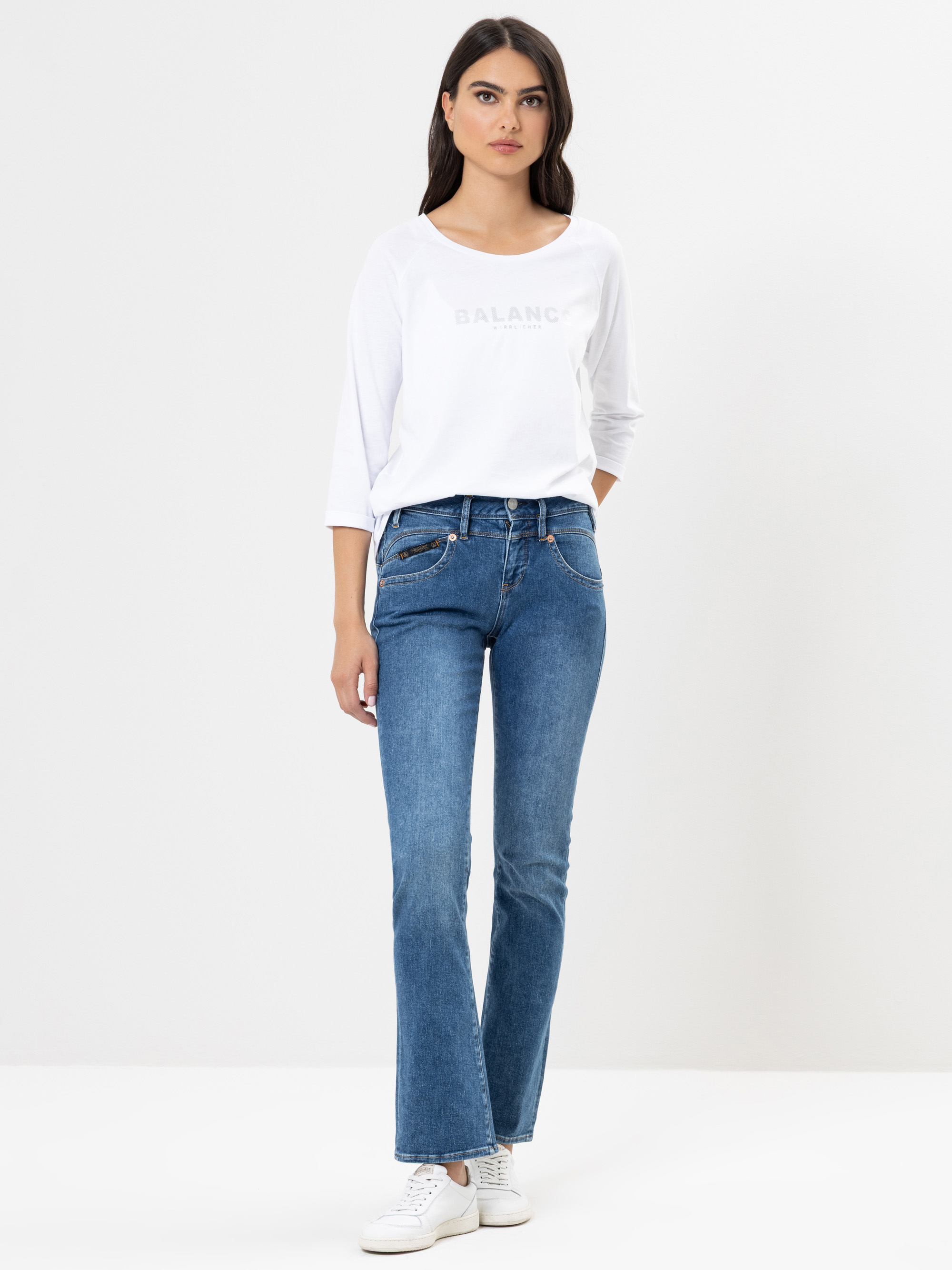 Eine Frau steht mit dem Gesicht nach vorne, trägt ein weißes langärmliges Hemd, blaue Bootcut-Jeans und weiße Turnschuhe vor einem schlichten weißen Hintergrund.