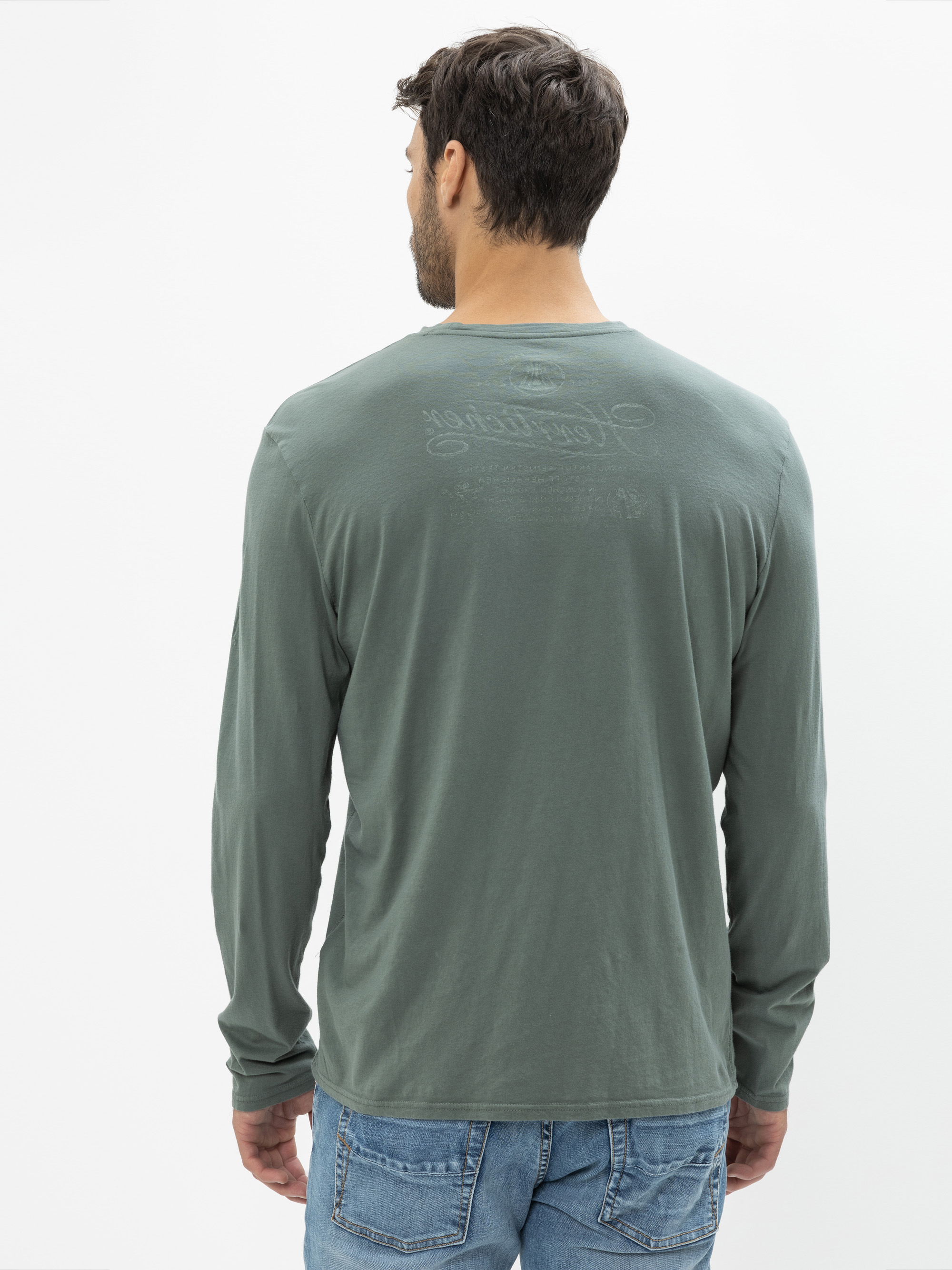 Ein Mann mit kurzen braunen Haaren steht in einem langärmeligen grünen Hemd und einer blauen Jeans und schaut von der Kamera weg. Der Hintergrund ist einfarbig weiß.