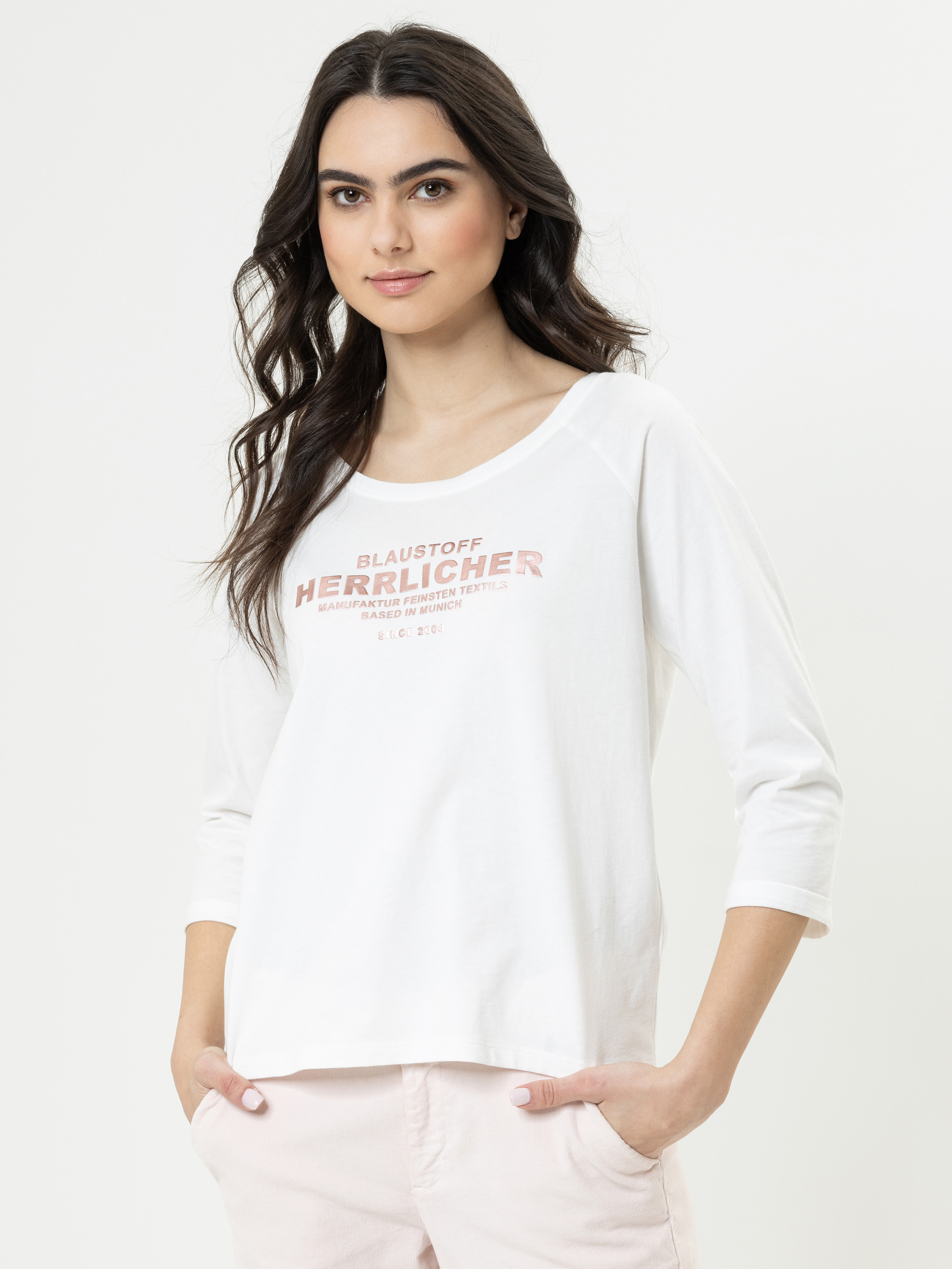 Eine Frau mit langen dunklen Haaren trägt ein weißes langärmeliges Hemd mit rosa Aufschrift und eine helle Hose. Sie steht mit den Händen in den Taschen, blickt nach vorne und lächelt leicht vor einem schlichten Hintergrund.