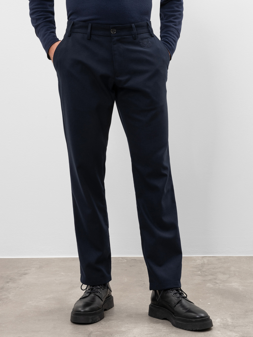 Eine Person steht mit den Händen in den Taschen, trägt eine dunkle marineblaue Anzughose, ein dunkles langärmeliges Hemd und schwarze Schnürschuhe vor einem schlichten hellen Hintergrund.