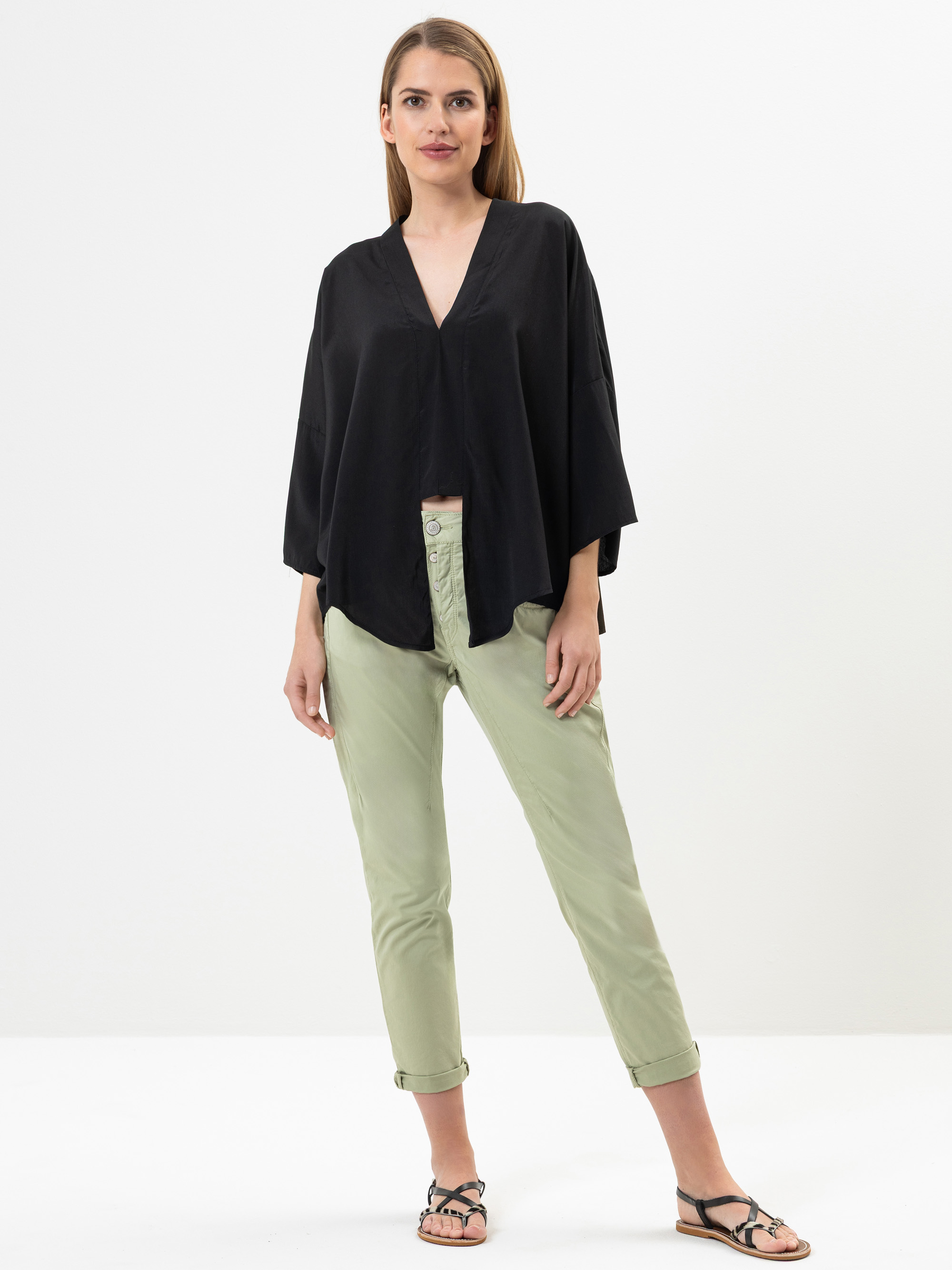Eine Frau steht vor einem schlichten Hintergrund und trägt ein lockeres schwarzes Oberteil mit V-Ausschnitt, eine hellgrüne Hose mit Bündchen und schwarze Sandalen. Sie hat glattes, hellbraunes Haar und eine entspannte, selbstbewusste Körperhaltung.