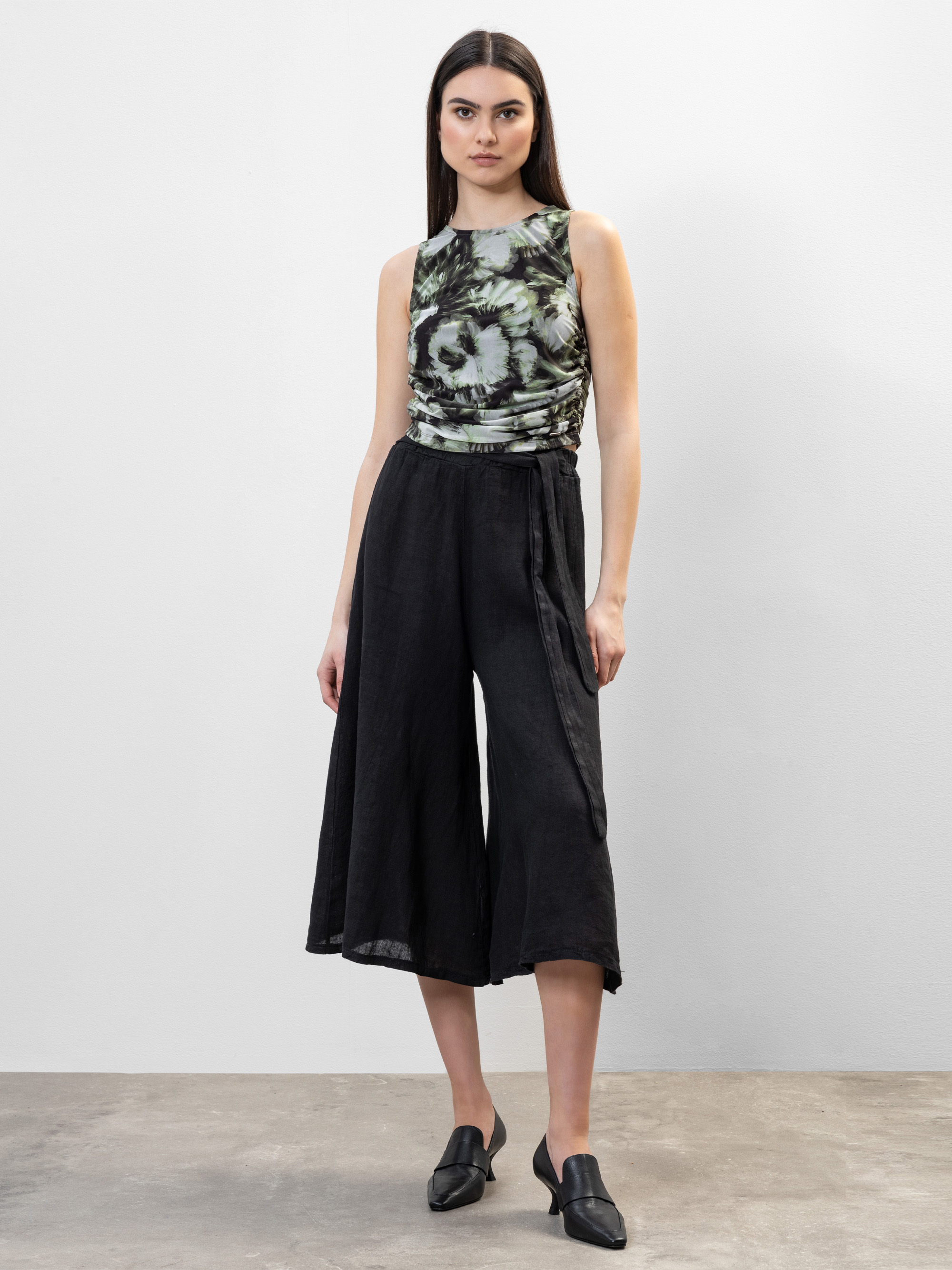 Eine Frau steht vor einer schlichten hellen Wand und trägt ein ärmelloses, abstrakt gemustertes Oberteil in Grün und Schwarz, eine schwarze Culottes mit weitem Bein und schwarze spitze Schuhe. Sie hat glattes dunkles Haar und einen neutralen Gesichtsau