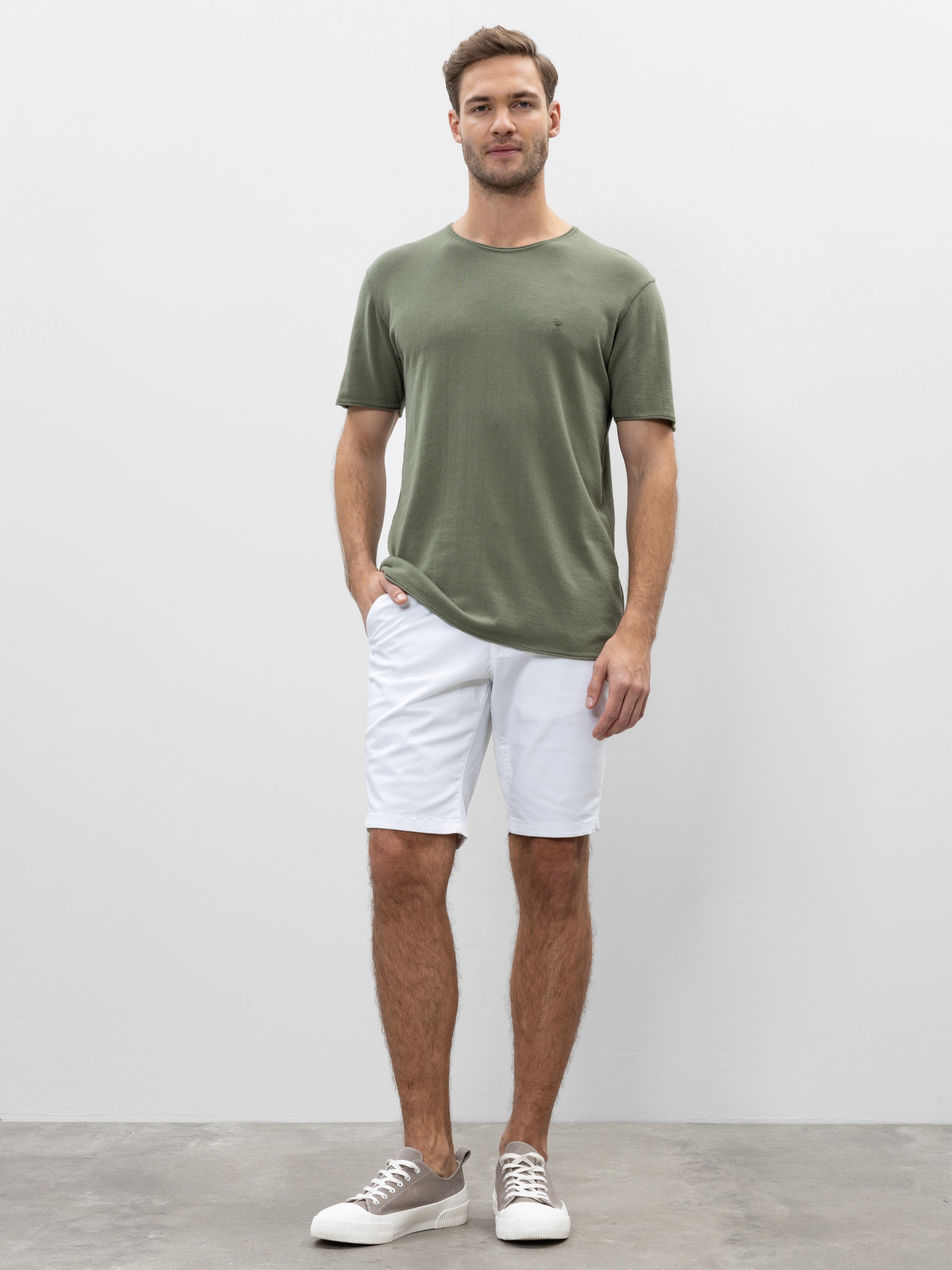 Ein Mann steht vor einem schlichten hellen Hintergrund und trägt ein olivgrünes kurzärmeliges T-Shirt, weiße Shorts und graue Turnschuhe. Er hat eine Hand in seiner Tasche und lächelt leicht.