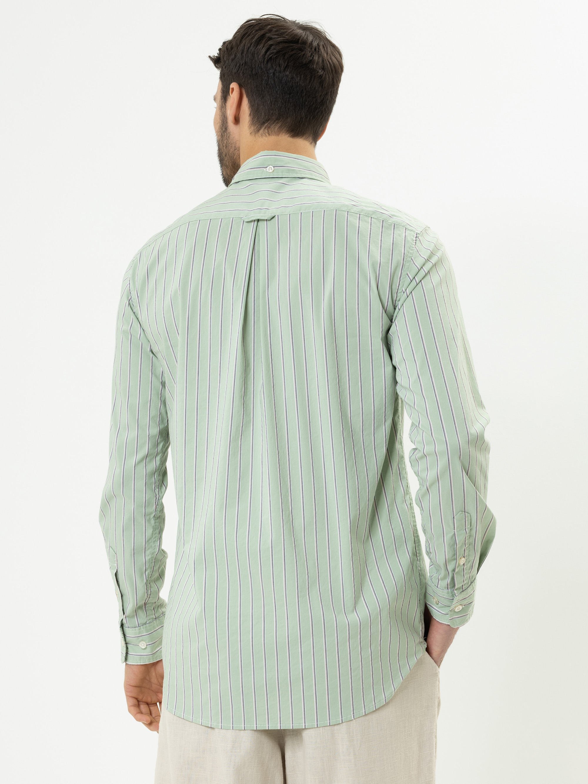 Ein Mann mit kurzen dunklen Haaren ist von hinten zu sehen. Er trägt ein hellgrünes, langärmeliges, geknöpftes Hemd mit dünnen vertikalen Streifen und eine helle Hose vor einem schlichten weißen Hintergrund.