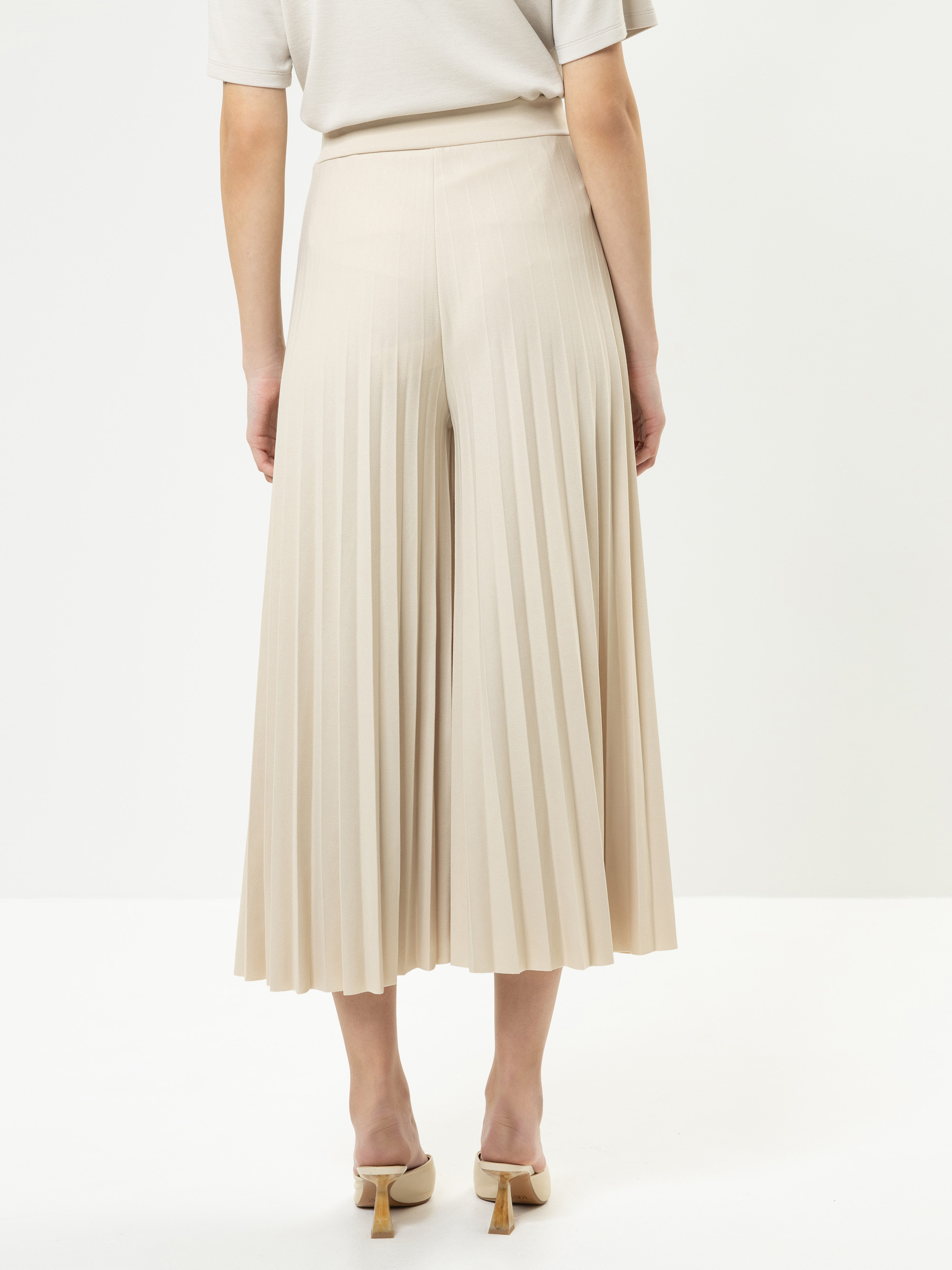 Eine Person steht mit dem Gesicht zur Wand und trägt einen beigen plissierten Midirock, ein passendes kurzärmeliges Oberteil und beigefarbene Schuhe mit Absatz. Der Hintergrund ist einfarbig weiß.