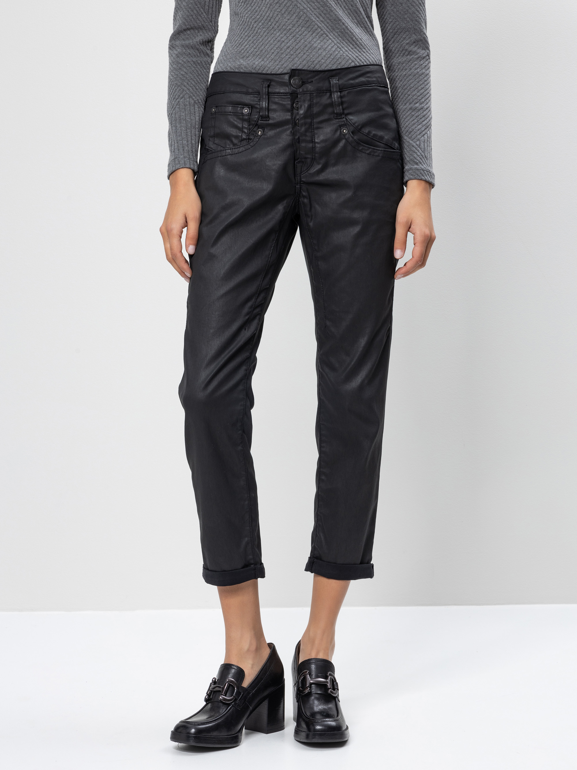 Eine Person, die eine schwarze, abgeschnittene Hose mit geradem Bein, ein graues, langärmeliges Oberteil und schwarze, hochhackige Slipper trägt, steht vor einem schlichten weißen Hintergrund. Das Bild ist an den Schultern abgeschnitten.