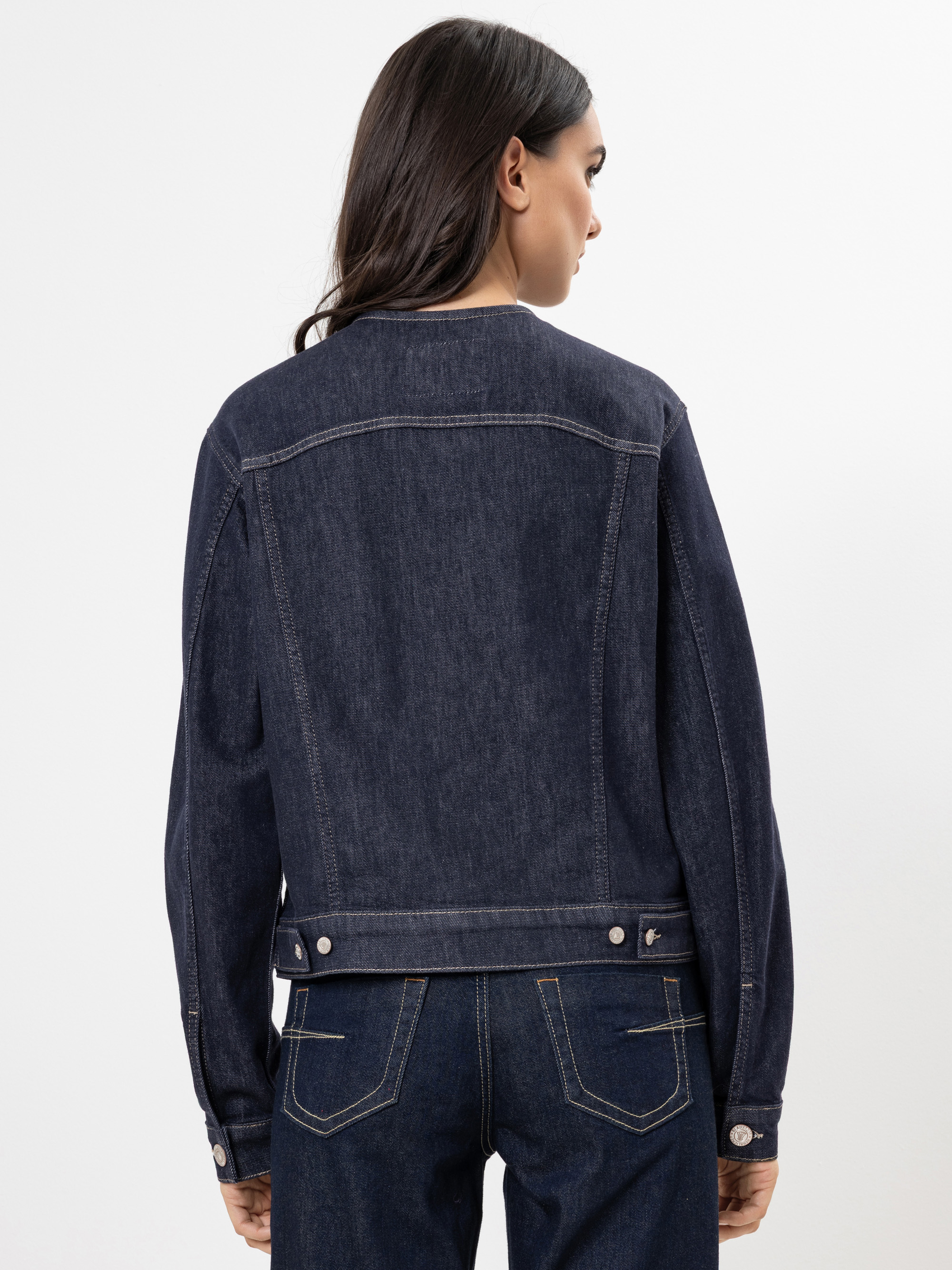 Eine Frau mit langen dunklen Haaren trägt eine dunkelblaue Jeansjacke und passende Jeans und steht mit dem Rücken zur Kamera vor einem schlichten weißen Hintergrund.